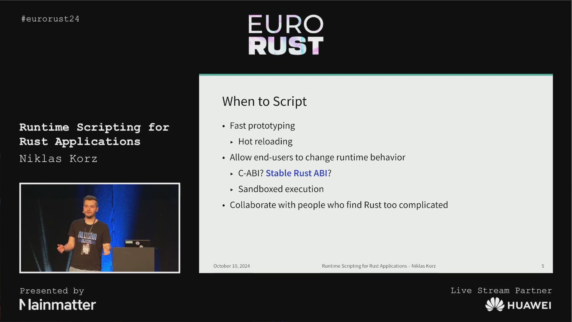 Niklas standing on the stage, presenting his talk "Runtime Scripting for Rust Applications". The slide shown next to him mentions reasons for integrating scripting languages into Rust applications: Fast prototyping (aided by hot reloading), extending applications without needing to alter their source code (through plugins), and to collaborate with people that find Rust too complicated.