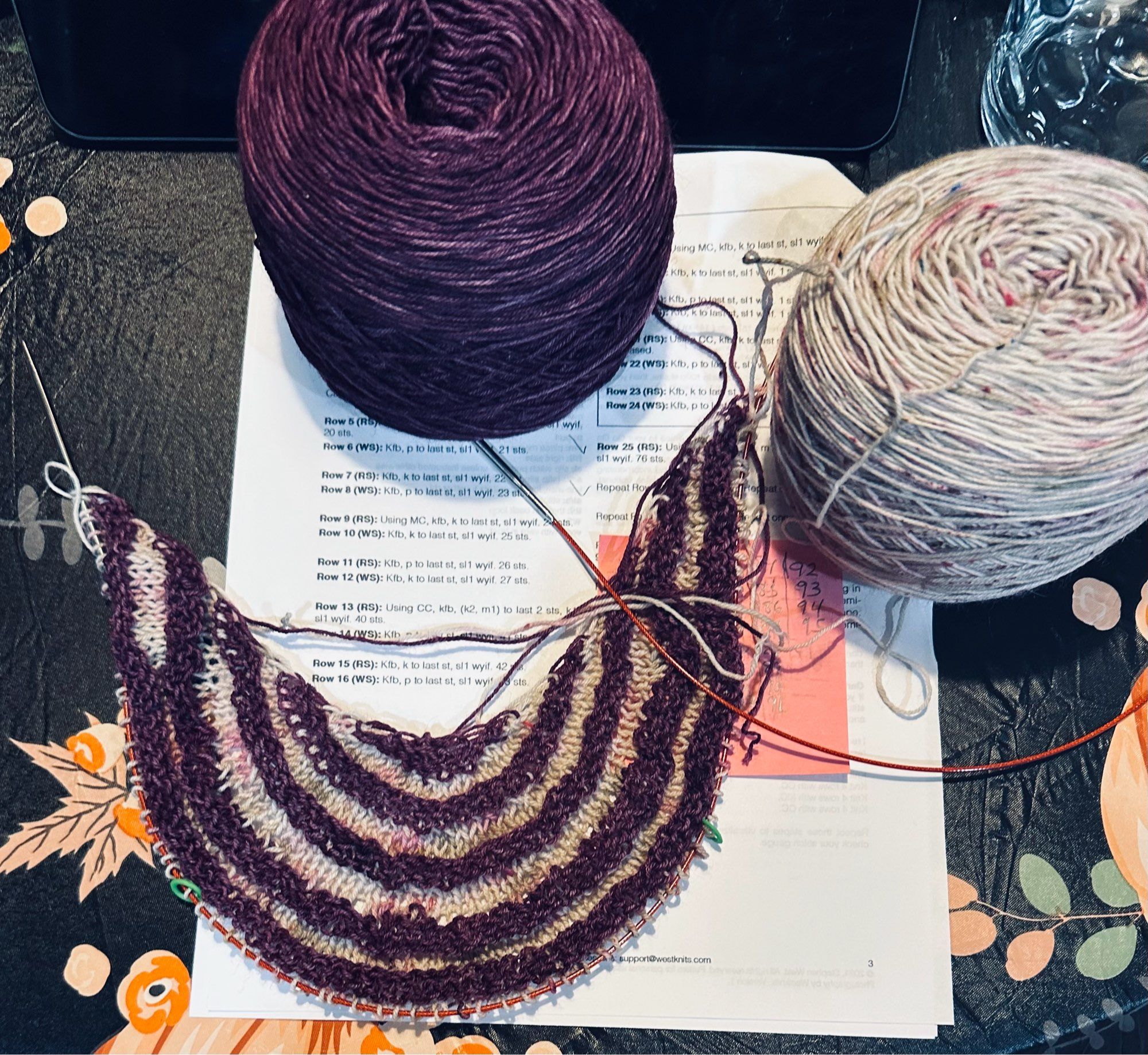 A striped semi-circular pattern in a maroon/purple yarn with a lighter pink-speckled contrast color is lying on top of a pattern on a black-fall themed tabletop.