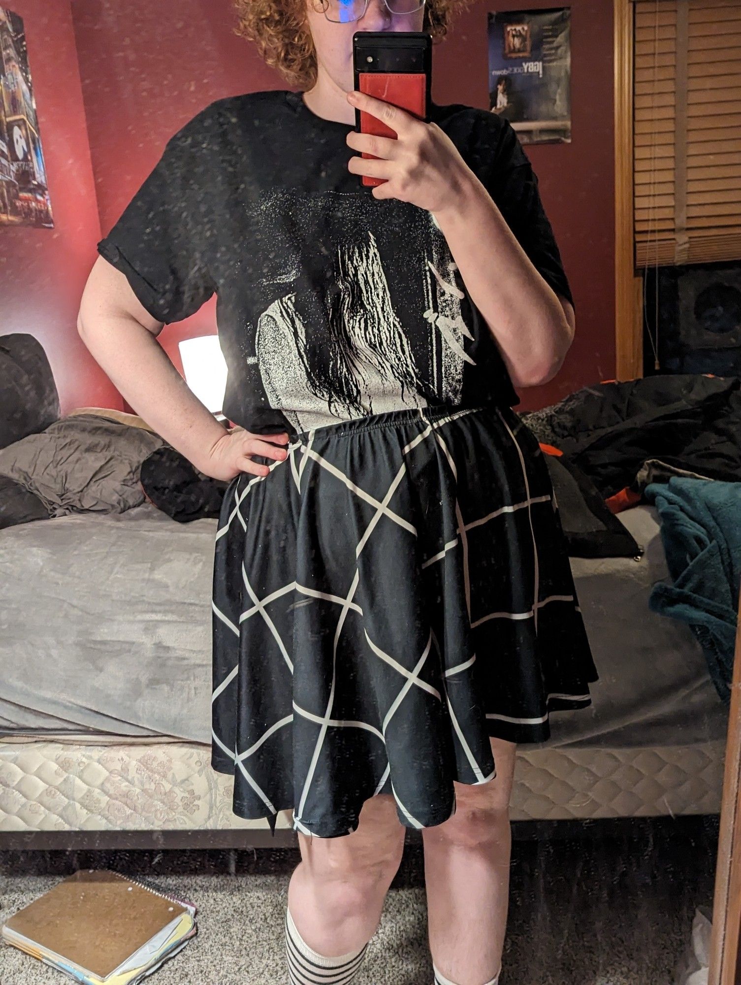 A chubby nonbinary person with red curly hair. They're wearing a black skirt with a white grid pattern, black and white striped knee socks, and a shirt with Sadako from Ringu on it.