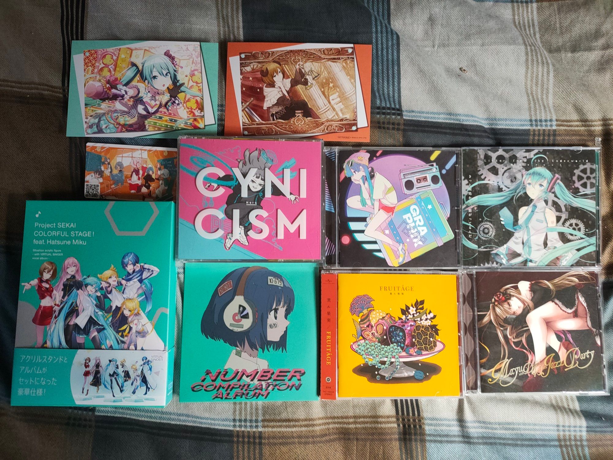 Photo of many albums.
On the far left is a dvd box for the Project Sekai Colorful Stage feat Hatsune Miku album which usually comes with acrylic stands but is just the album box. 
To the right of that is the album Cynicism by Neru on top and Number one Compilation album on bottom. The card for the later album is also next to Cynicism above the Project Sekai box and above both of those are two cards featuring Miku and Meiko from Project Sekai.
To the right of that is Graphix by HachioujiP on top and Fruitage by NILFRUITS on the bottom.
Lastly to the far right is Linker to the Underworld on top and Mayu Jazz Party on the bottom.