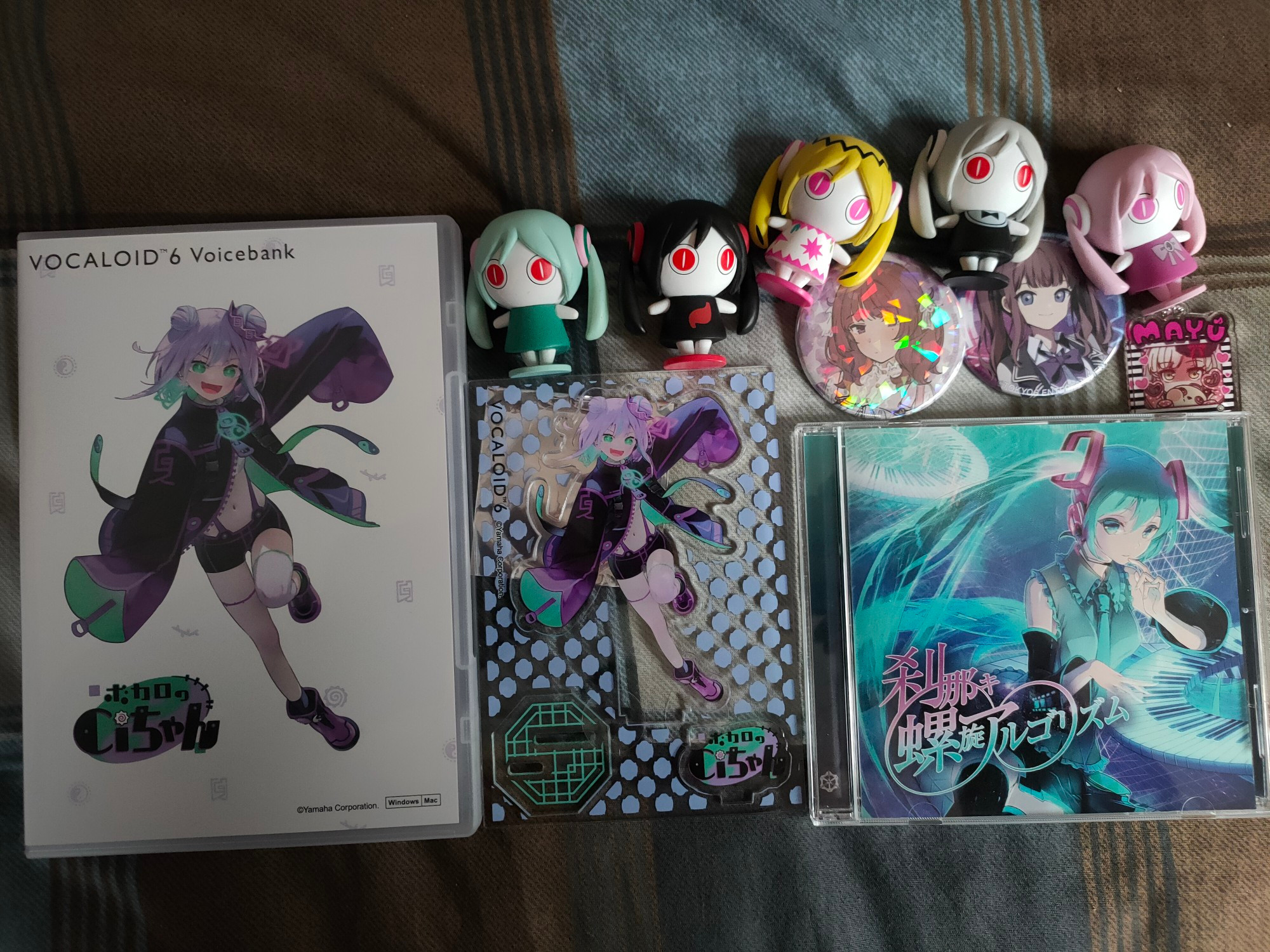 Picture of merchandise on a bed.
On the far left is a DVD case for VOCALOID 6 Ci-chan with the acrylic stand she came with to the right of her.
Next to the stand is the album Setsunaki∞Rasen Algorithm with Hatsune Miku on the cover and features songs from both her and Ring Suzune.
On the top of the CD are 2 badges of Natsuki Karin and a small Mayu keychain.
Going across the top of that and the acrylic stand are 5 Aimaina figures, a complete set from the first round of gachapon.