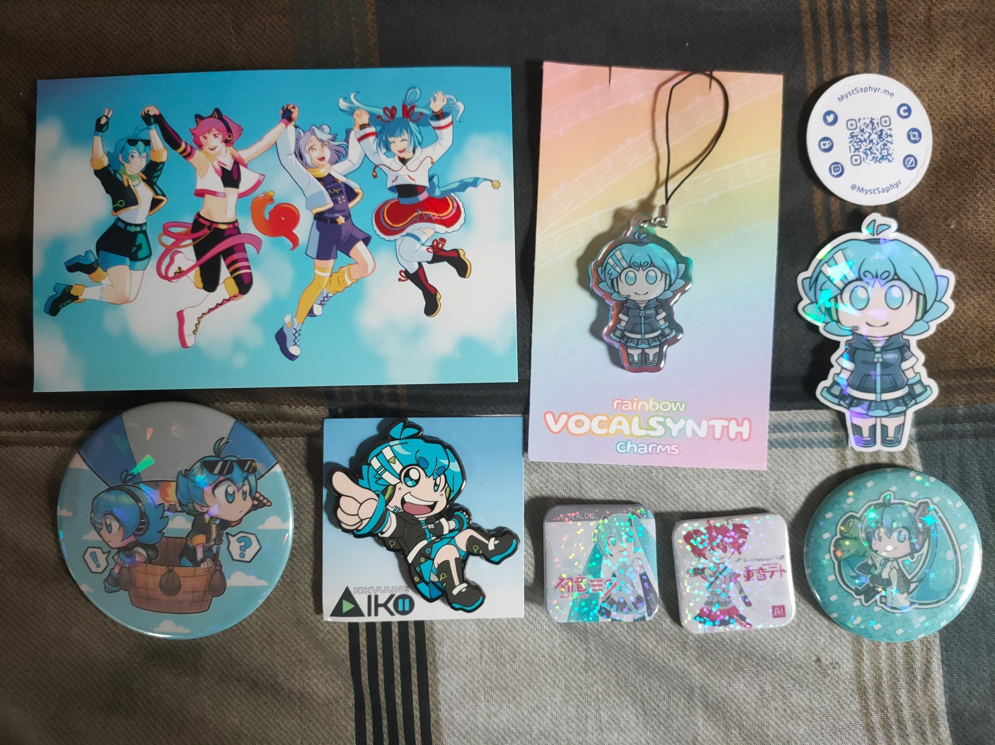 A collection of merch on a patterned bedsheet. 

On the top left is a postcard featuring 4 vocals from the synth engine "Mikoto Studio".

On the bottom left corner is a giant pin of Kikyuune Aiko and Kikyuune Aiko 3510 in a hot air balloon. Next to it on the right is a metal pin of Kikyuune Aiko.

In the bottom right corner is a badge of Hatsune Miku holding a leek plush. To the left of it are 2 square badges, one of Hatsune Miku in the style of her Vocaloid 2 box and one of Kasane Teto in the style of her Synthesizer V box.

On the top right is a qr code on a coin to take you to MystSaphyrs sites. Bellow that is a sticker of Kikyuune Aiko. To the left of those is a charm of Kikyuune Aiko in the same style as the sticker.