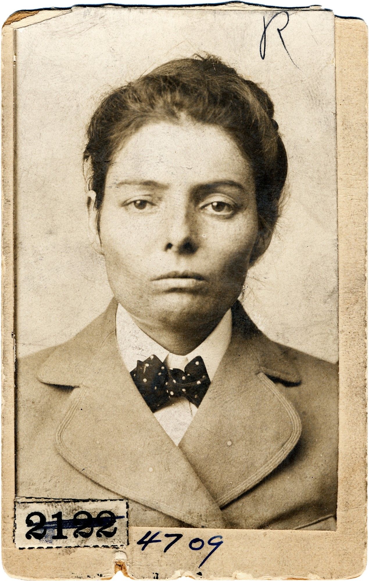 black and white photo of a stern and powerful woman with dark hair and dark eyes looking directly into the camera, wearing a coat and white shirt with a small polka dot bowtie. the number 2122 is stamped on the photo but crossed out, with the number 4709 written in ink next to that