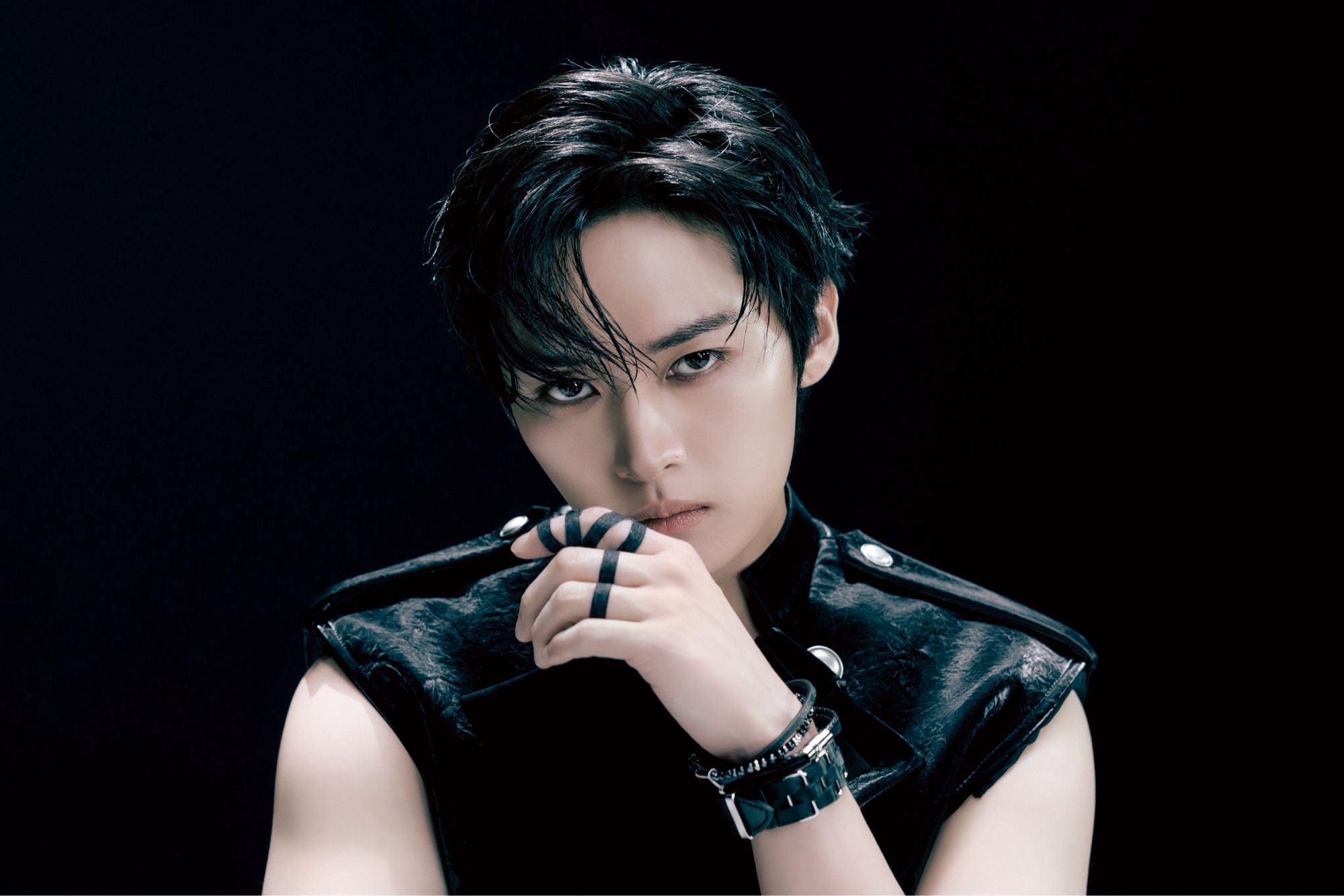 Lee Know in front of a dark background. He’s glaring at the camera wearing a black vest with multiple black bracelets/accessories.