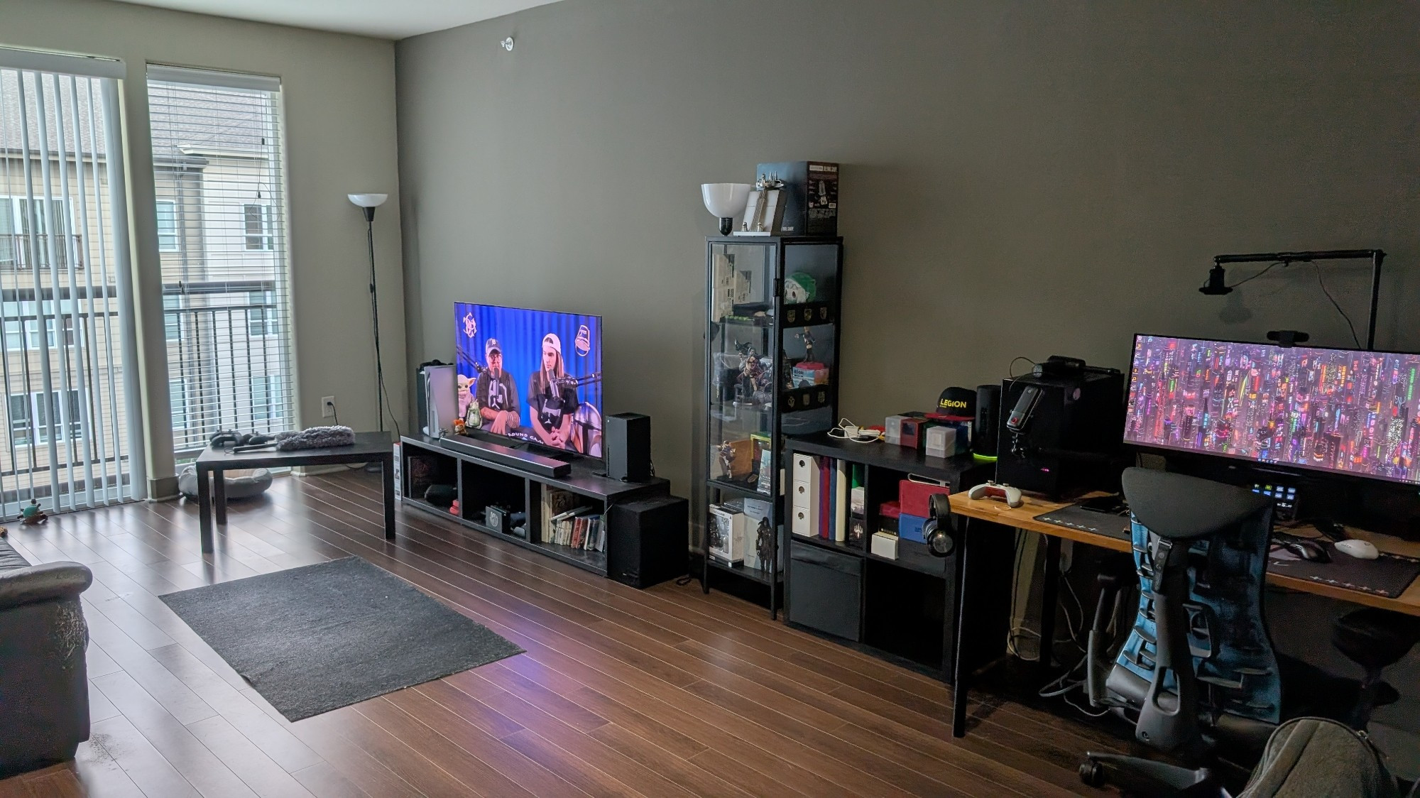 Living room area with a TV showing a stream, a tall glass display case filled with nerd trinkets, a small box shelf filled with card game storage, and a desk with a small computer and an ultra wide monitor, all of which along a wall.