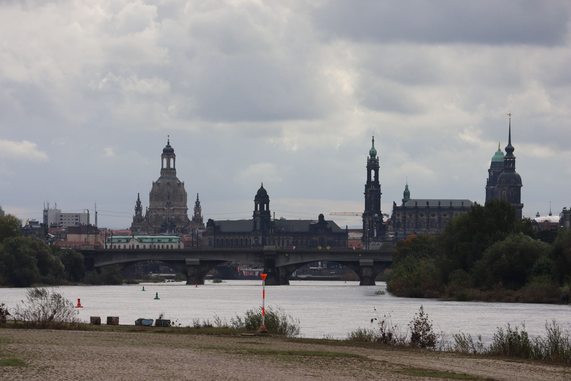 Blick auf die Dresdner Altstadt vom Elbeufer aus, im Vordergrund ein Stück Elbestrand. Der Himmel darüber ist grau-bewölkt, die Sonne lässt sich hinter den Wolken jedoch erahnen.