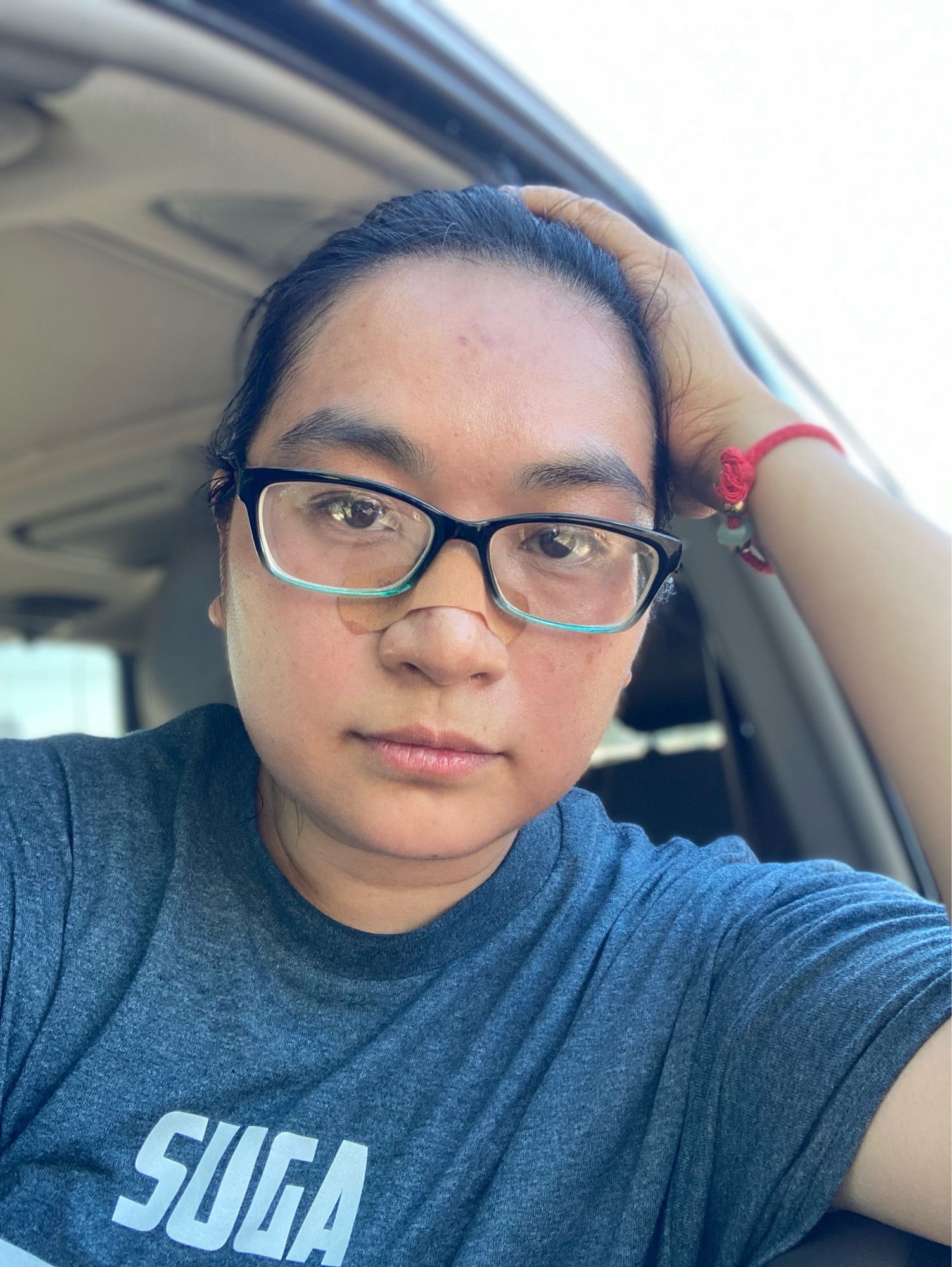 Asian nonbinary person wearing plastic rimmed glasses, a SUGA 93 T-shirt, and a red string + gold bead and green stone bracelet, leaning out an open car window on one elbow, hand on head. There is a bandaid across the bridge of their nose under eh glasses.