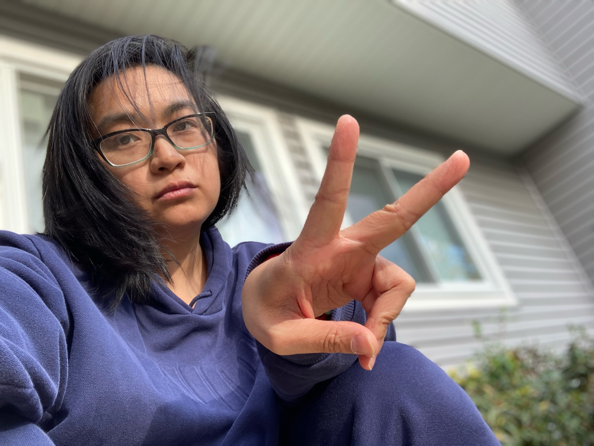 Photo of Asian nonbinary person with glasses on wearing a navy sweat suit combo outside in front of a house, doing a peace sign
