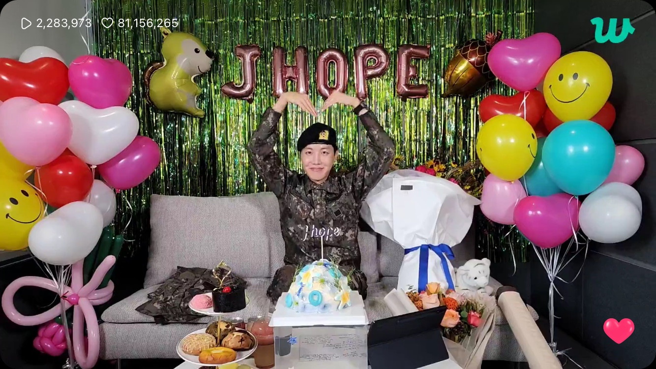 J-Hope of BTS in military uniform making a large heart shape with his hands over his head. He is surrounded by balloons and decorations and flower bouquets, cake and snacks on a table in front of him