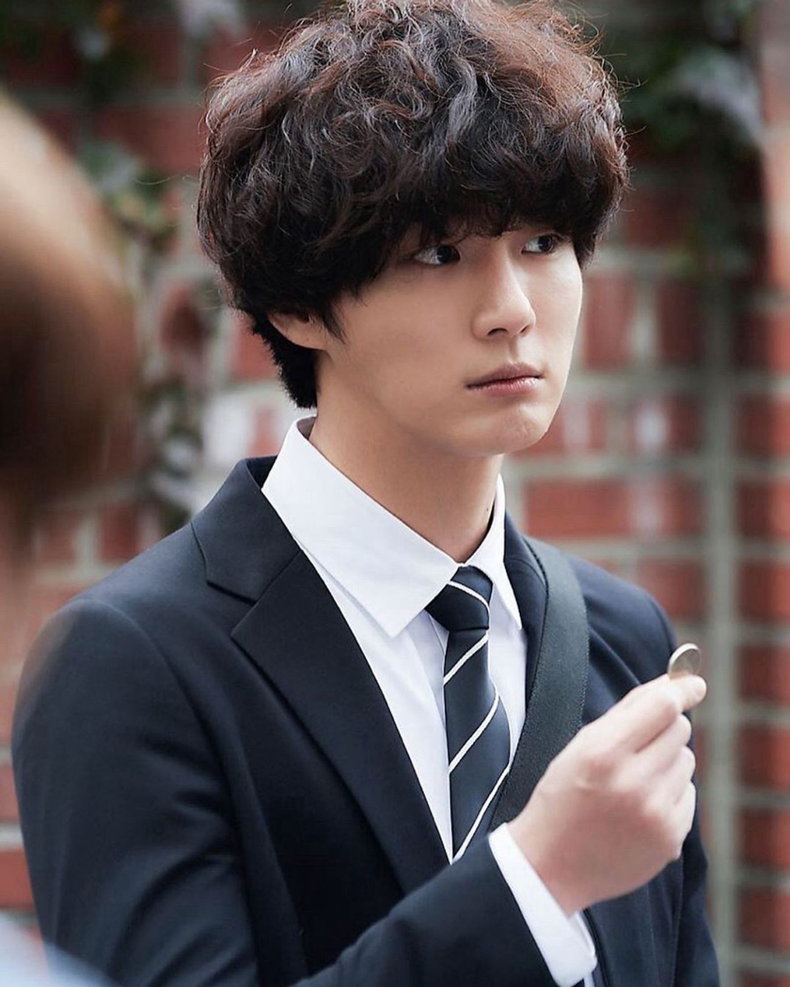 Yoon ShoYoon playing Yook DongSik in “Psychopath Diary” …An Asian man with fluffy hair in a business suit, holding up a coin and looking pensively off into the distance