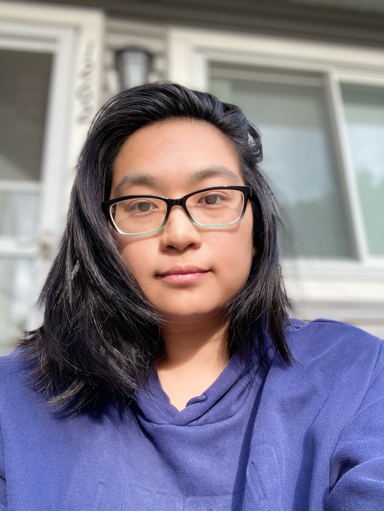 Photo of Asian nonbinary person with glasses on wearing a navy sweat suit combo sitting outside in front of a house
