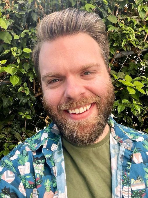 Selfie of Robo outdoors in front of a leaf covered fence. He wears a green shirt with a blue button up shirt worn open over it. 