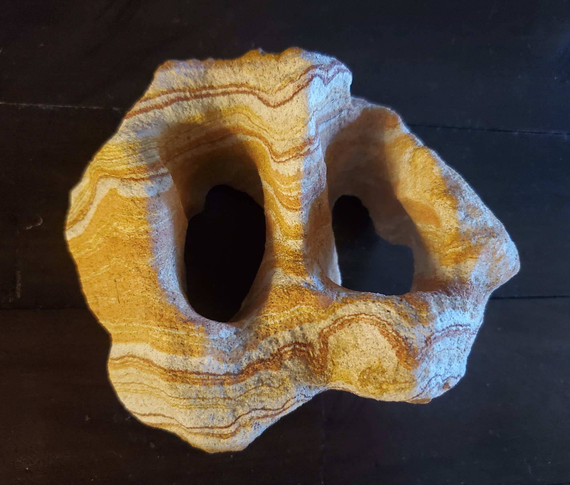 A hand sized chunk of sandstone with two large holes through it. It has lots of straiations and waves in various oranges and whites.