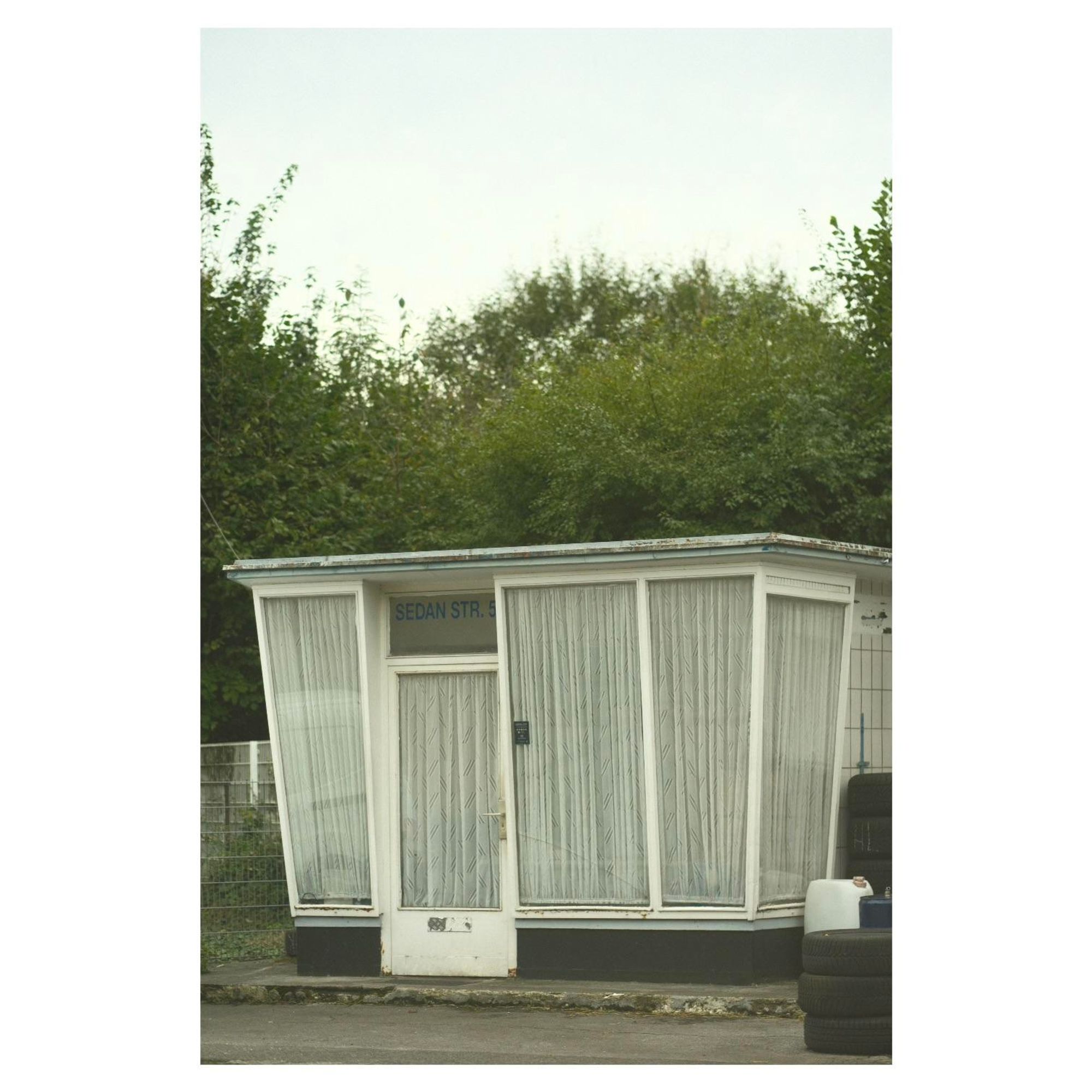 Photo. Außenansicht eines kleinen Büros einer Autowerkstatt aus den 60er Jahren. Schräge, hohe Fenster im Stile der 60er umlaufen das einstöckige Gebäude und reichen fast bis auf den Boden. Die Fenster sind von innen komplett mit weißen Vorhängen bedeckt. 