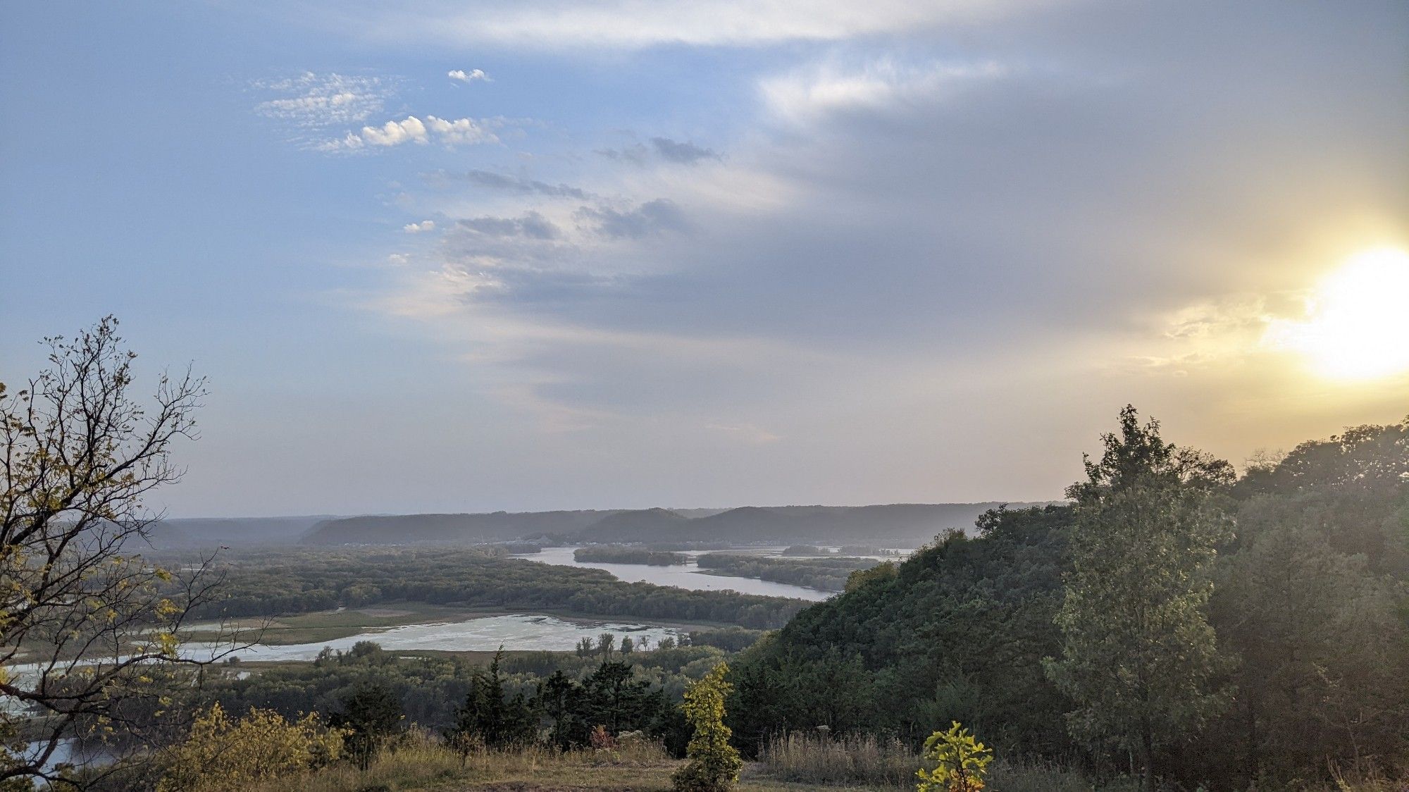 Hazy Mississippi river valley. One last fishing boat on the main channel making for home. Sun a bog hazy blob on the right hand side of the photo. On the left, the favorite tree of an indigo bunting, where it perches at sunset and start singing its song.