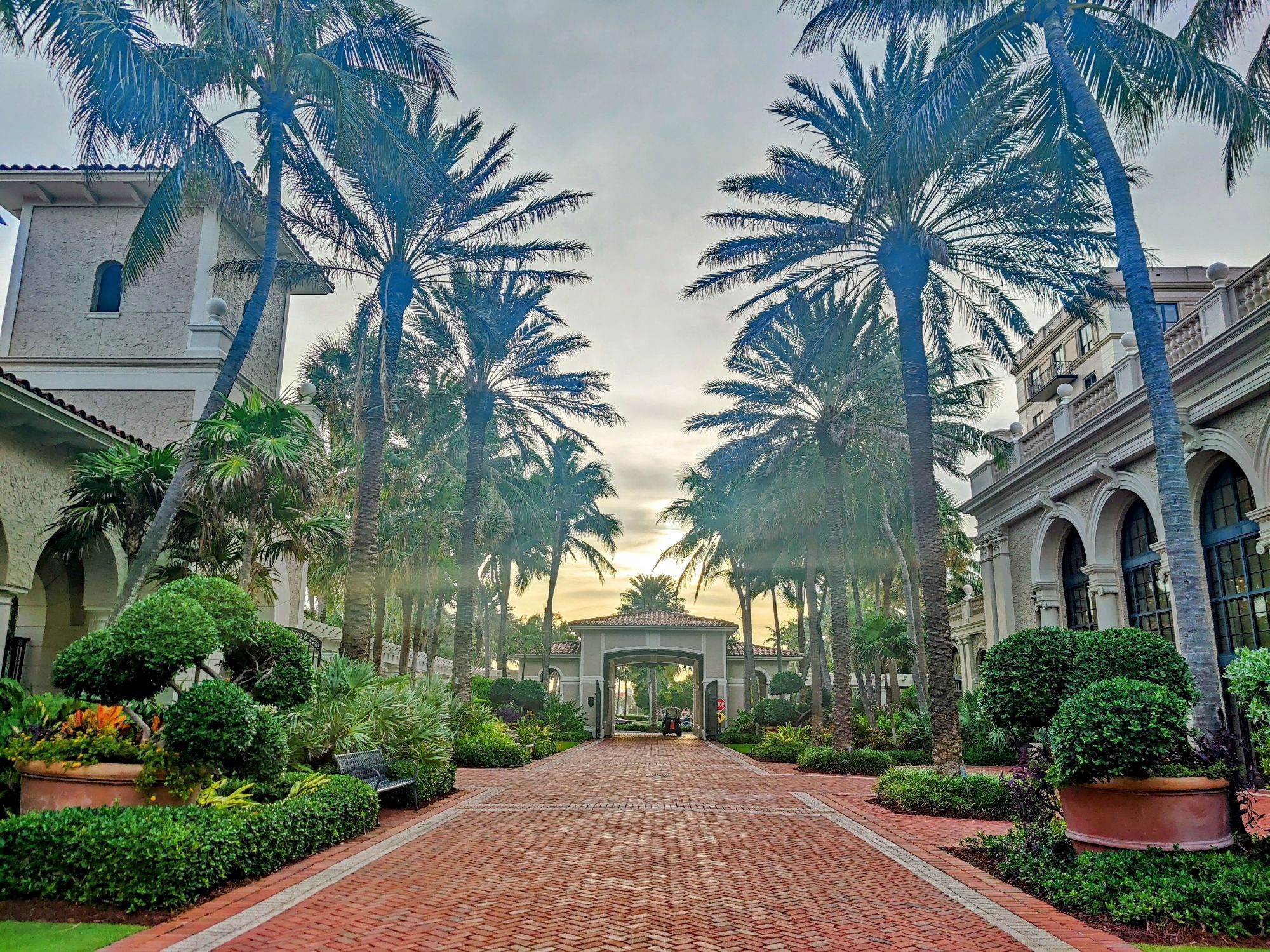 A red brick, cobblestone road recedes into the distance and a stuccoed, terra cotta roofed gate. The road is flanked by two stuccoed, two storied buildings separated by lots of green foliage and tall palm trees.