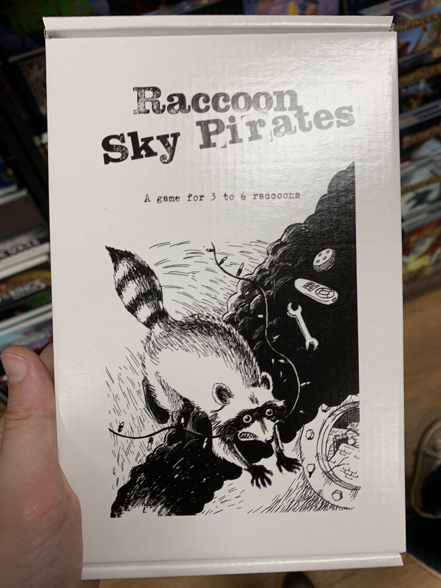 Raccoon Sky Pirates, a game for 3 to 6 raccoons. A white box with a drawing of a raccoon leaping from an explosion with a dangled line of Christmas lights in its mouth.