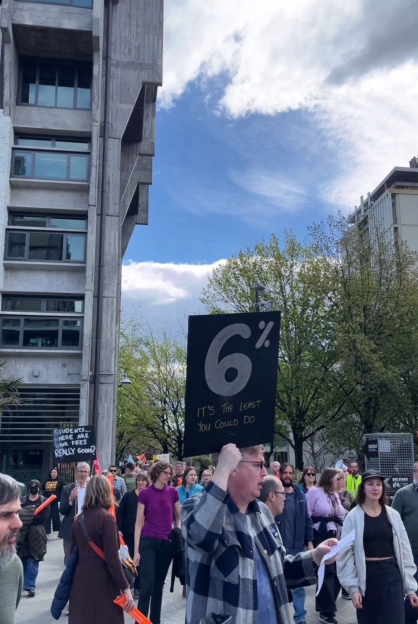 A crowd of hundreds of striking workers walk in a procession around the UC campus, past the concrete brutalist Matariki building with spring trees in the background.