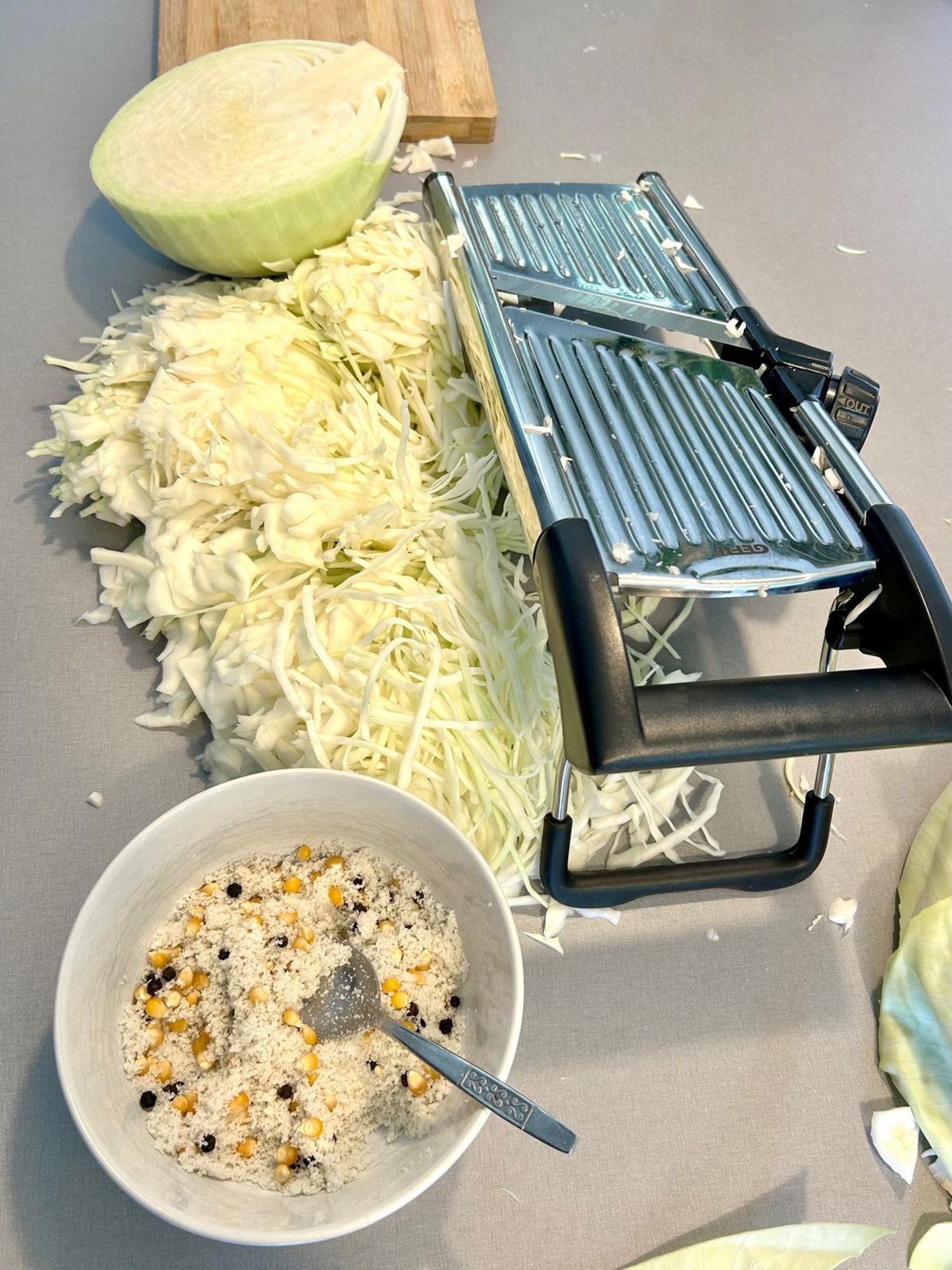 Kitchen counter top with half a cabbage, sliced cabbage, wooden chopping board, a mandoline, and a bowl full of salt, dried corn and peppercorns.