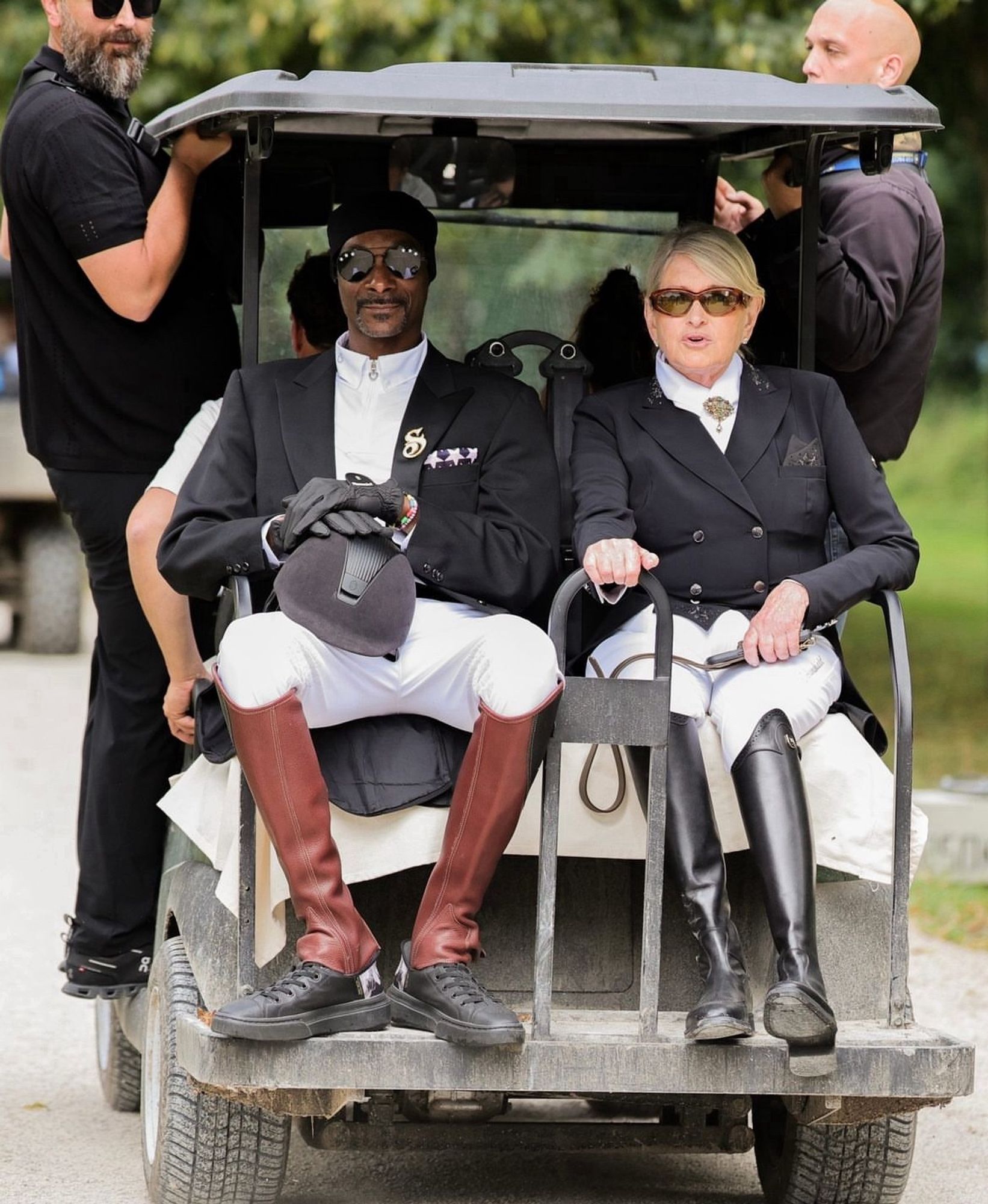 Snoop and Martha on the back of a golf cart facing out. They're dressed in high boots, white pants, and formal black jackets and sunglasses.