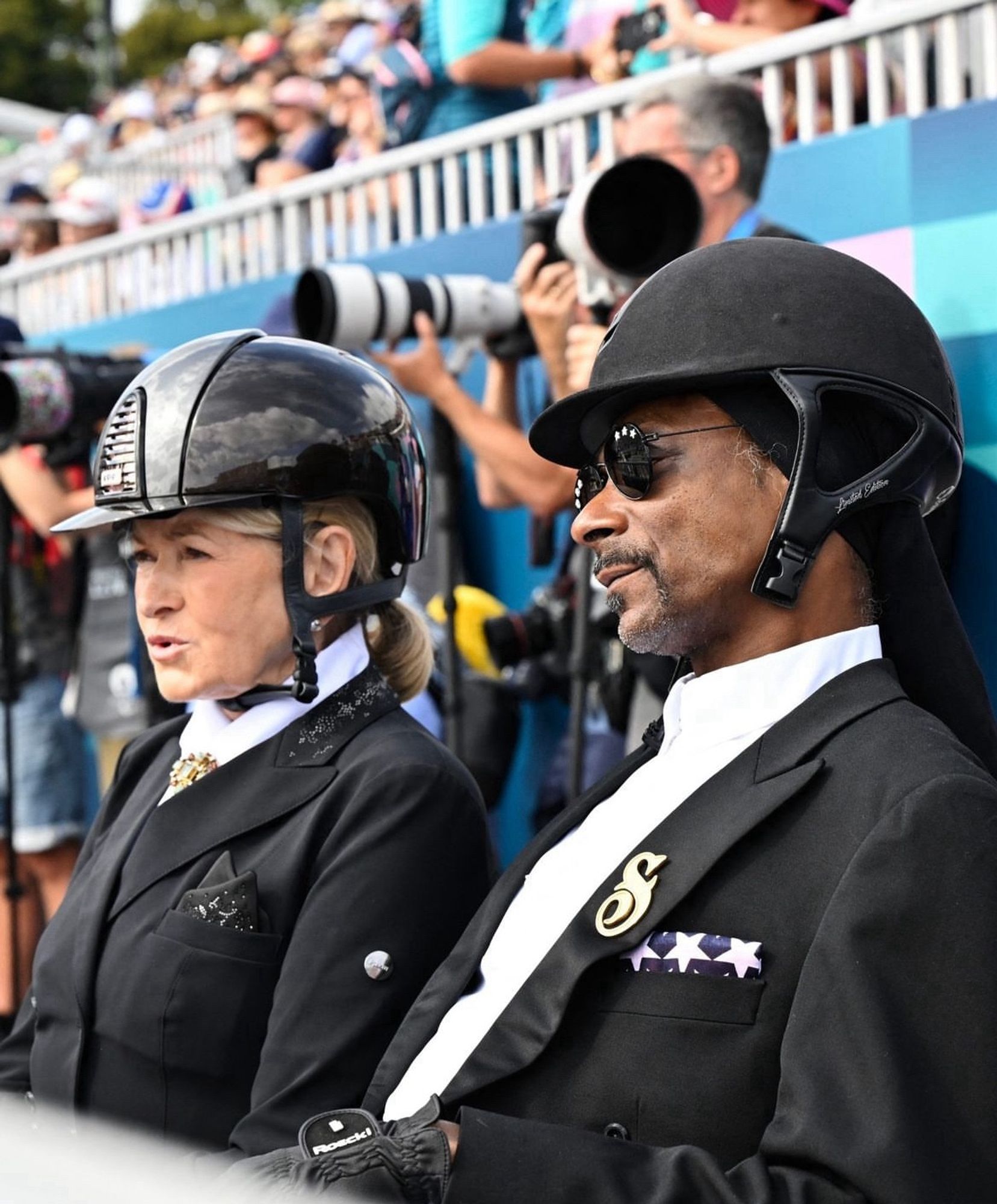 Snoop and Martha sitting next to each other at the dressage event. They're wearing formal black jwfkrye and white shirts. Snoop's jacket had a large gold "S" on it. They have horse riding helmets on.