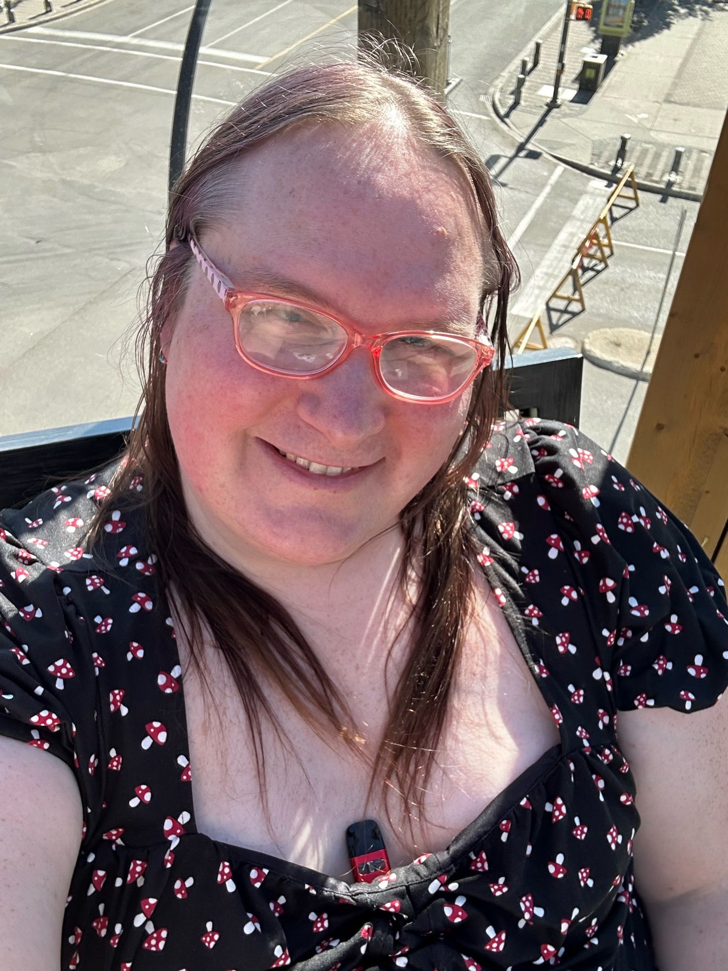 Me sitting on a rooftop patio, wearing a black dress with mushroom print and getting far too exposed to the hated daystar but still smiling like I’m having fun (I am!)