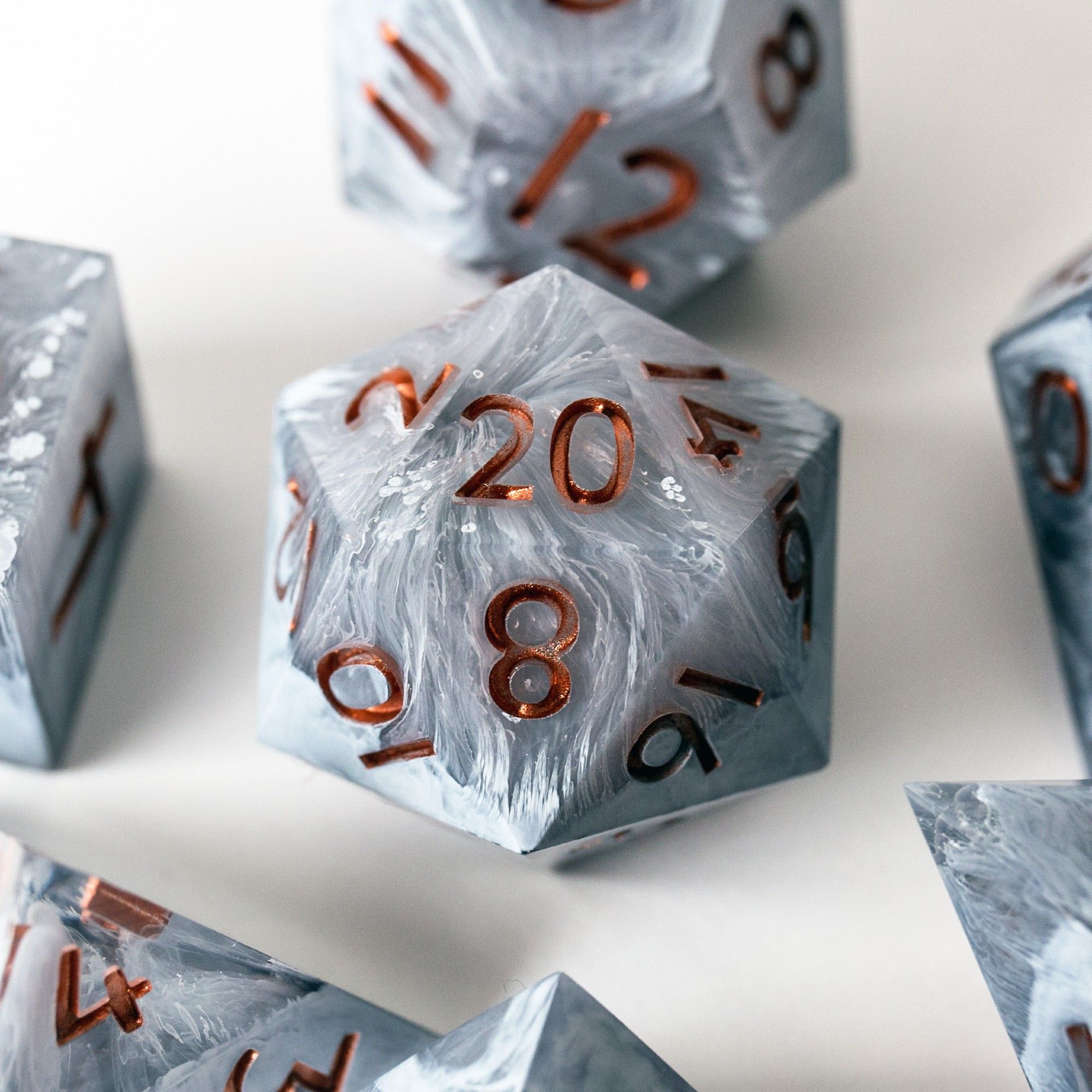 Eine Detailaufnahme des D20. Der Würfel ist grau und durch den Petri Dish Effekt ist er von weißen Verwirbelungen durchzogen. Die nummern sind in Kupfer gehalten. Der D20 liegt auf einem weißen Untergrund und ist von anderen Würfeln aus dem Set umgeben, die man aber nicht erkennen kann.