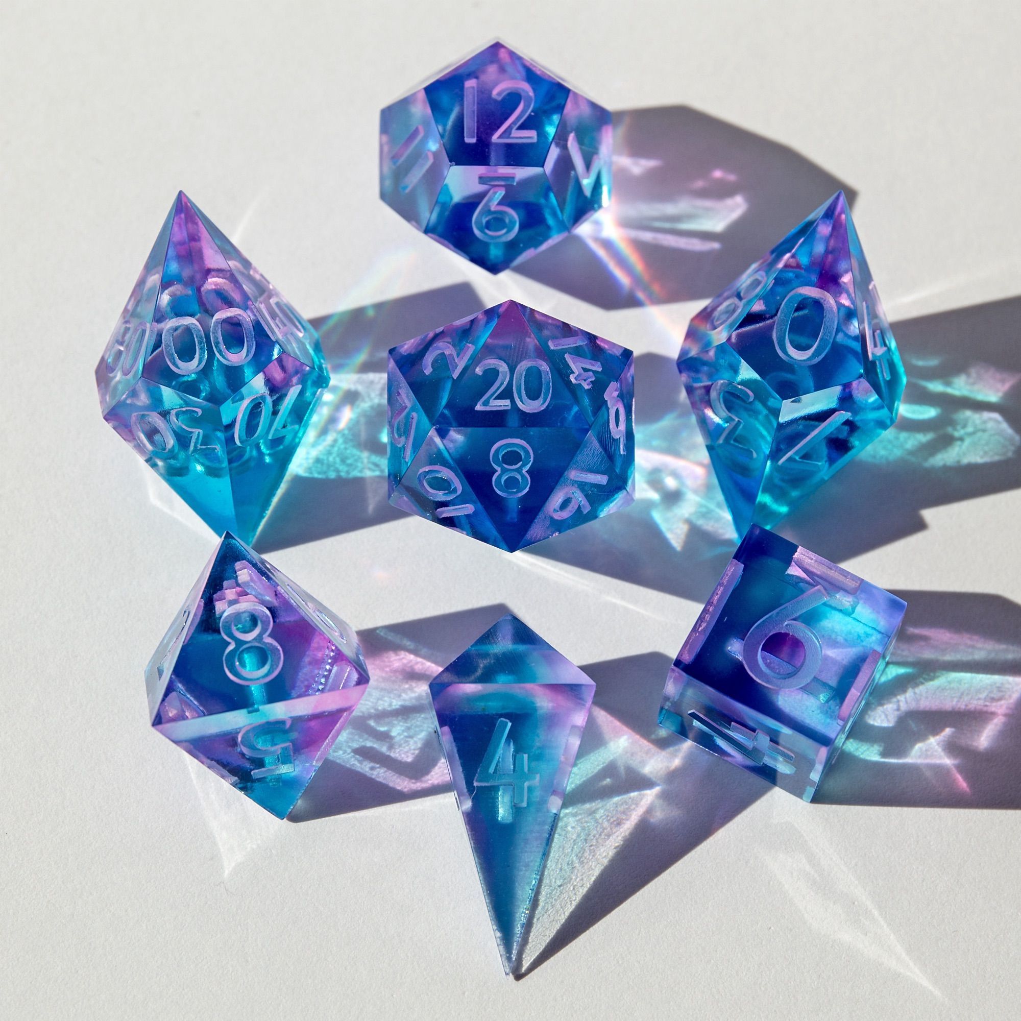 Hier ist das ganze blau/lila Würfelset zu sehen. Das Set besteht aus einem D4 Shard, D6, D8, D10, D00, D12 und D20. Das Set liegt auf einer weißen Unterlage und die Sonne erzeugt Reflektionen, welche auf dem Foto für einen tollen Effekt sorgen. Alle Würfel sind um den D20 herum angeordnet.