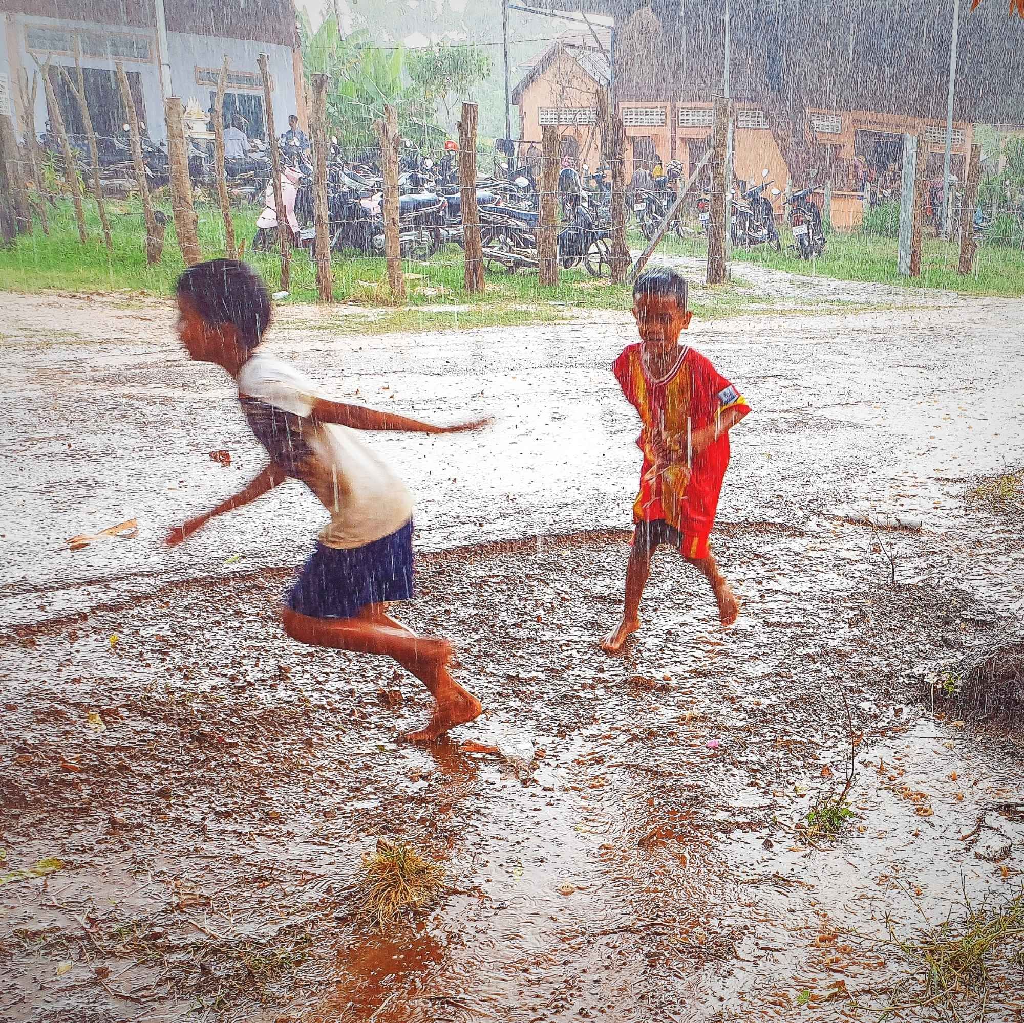 Photo shows 2 Khmer boys in t-shirt and shorts running and playing in a heavy downpour of rain. In the background there are some typical khmer houses, green grass and parked motorbikes. A roadside photo, the rain hitting the asphalt bounce back up by the force of the rain. The road shoulder is having a small stream of rainwater flowing toward the photographer off the road shoulder.