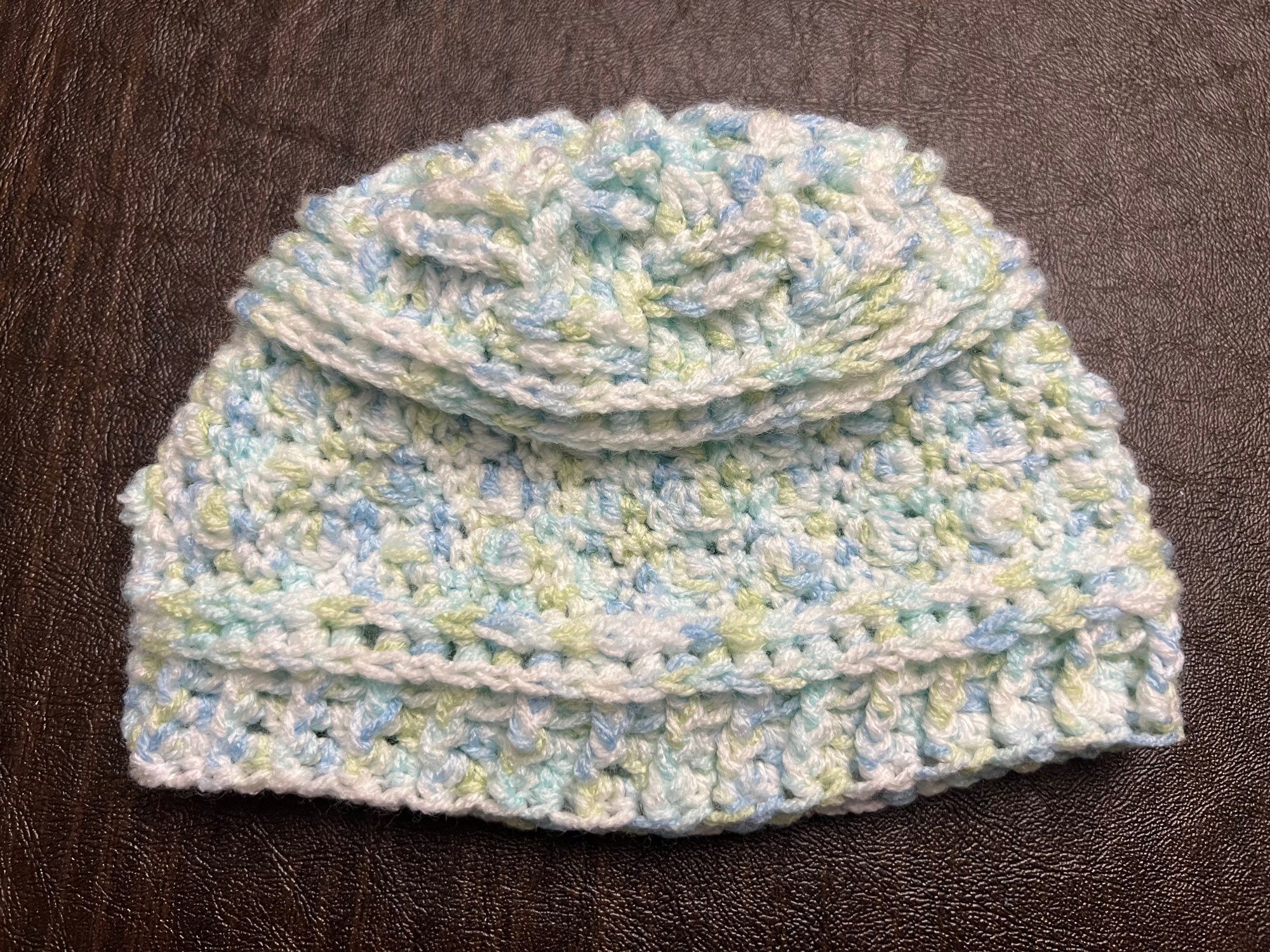 a photo of a baby hat crocheted from multi-colored yarn (white, light blue, light green) on a dark background