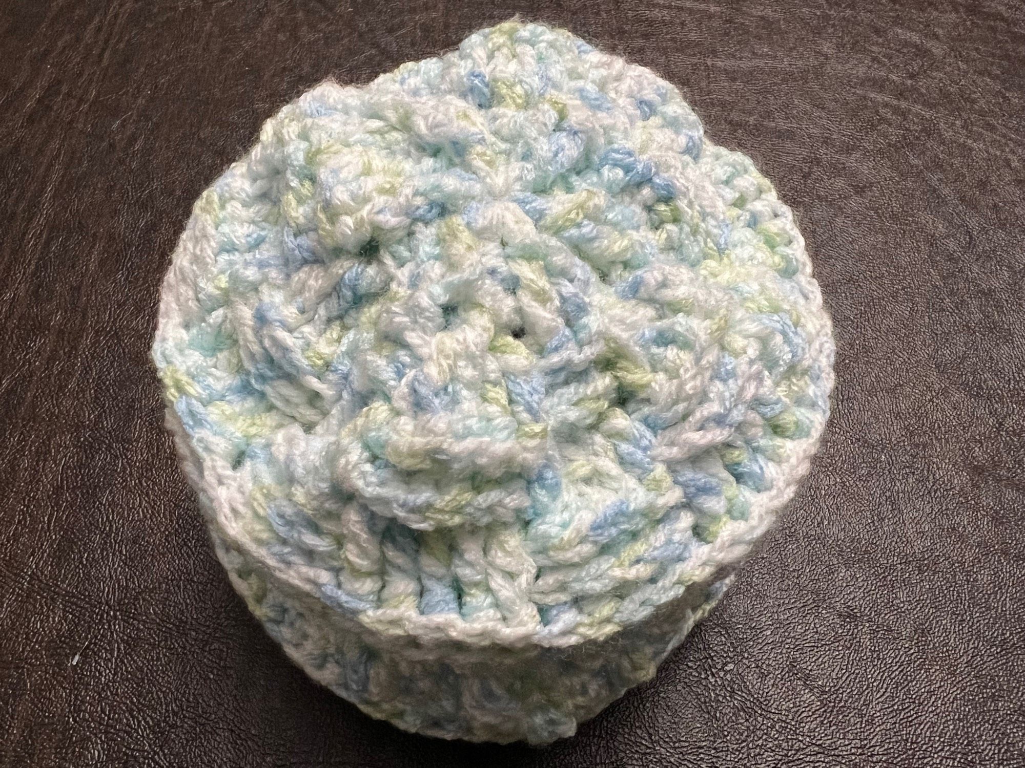 a top view photo of a baby hat crocheted from multi-colored yarn (white, light blue, light green) on a dark background