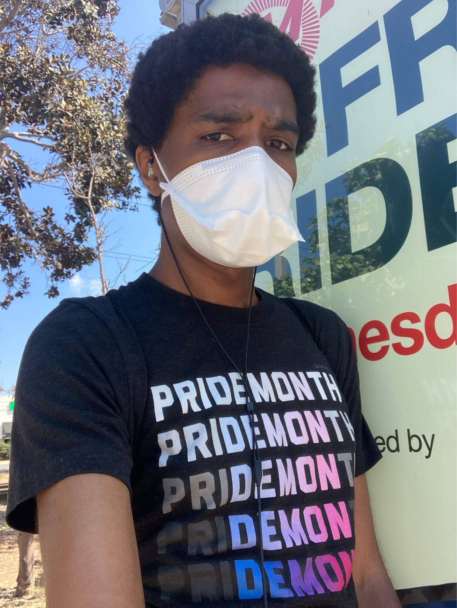 Vicki wears a t-shirt that reads “pridemonth”, the edge letter fading away to reveal the word “demon” in violet, blue and deep pink. She also wears a mask.