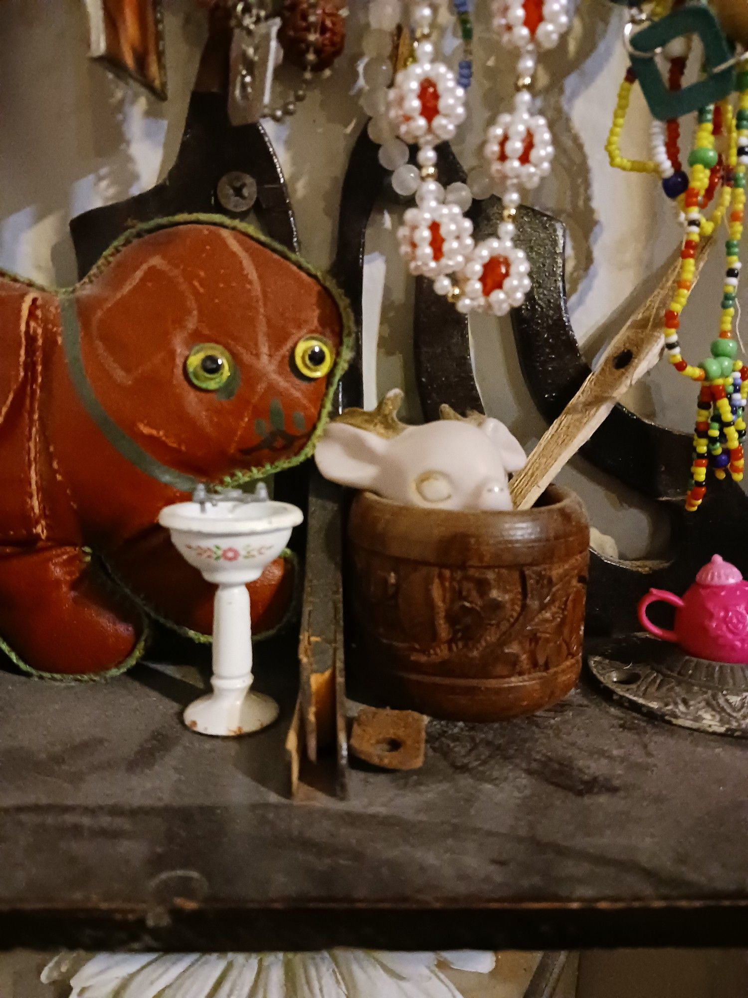 A bizarre red oil cloth child's purse shaped like a cat from the 1920s sits on a cut wood shelf next to a 2 inch tall cast aluminum sink. Beside that, a carved wood napkin ring holds the pink and gold head of a broken porcelain deer. A pink barbie teapot sits in an art nouveau doorknob plate, abd a barrier of necklaces hang above