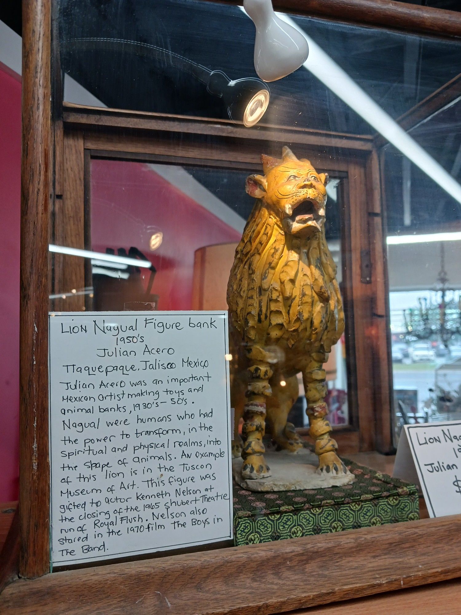 A glass case with a labeled sculptural piece of a Mexican carved wooden lion
The label reads
"Lion Nagual Figure bank
1950s
Julian Acero
TTaquepaque Jalisco Mexico
Julian Acero was an important Mexican artist making toys abs animal banks, 1930s-50s.
Nagual were humans who had the power to transform, in the spiritual and physical realms, into the shape of animals. An example of this lion is in the Tuscan Museum of Art. This figure was gifted to actor Kenneth Nelson at the closing of the 1965 Shubert Theater run of Royal Flush. Nelson also starred in the 1970 film Boys in the Band."