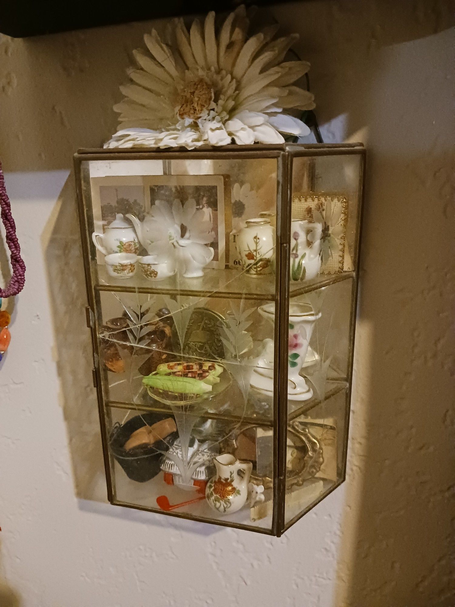 A cracked brass and etched glass cabinet, only about 9 inches tall, hands on my wall. On its 3 shelves are a variety of tiny goods, including a Dollhouse sized teaset from occupied Japan, 2 inch state photos from 1969, a tiny 1920s couple photo, Dollhouse food and dishes,a pinky nail sized mouse, tiny skates for a Patsy doll, corn in a glasses lens, abd a q9th century cast iron coal scuttle. A large fake daisy sits on top.