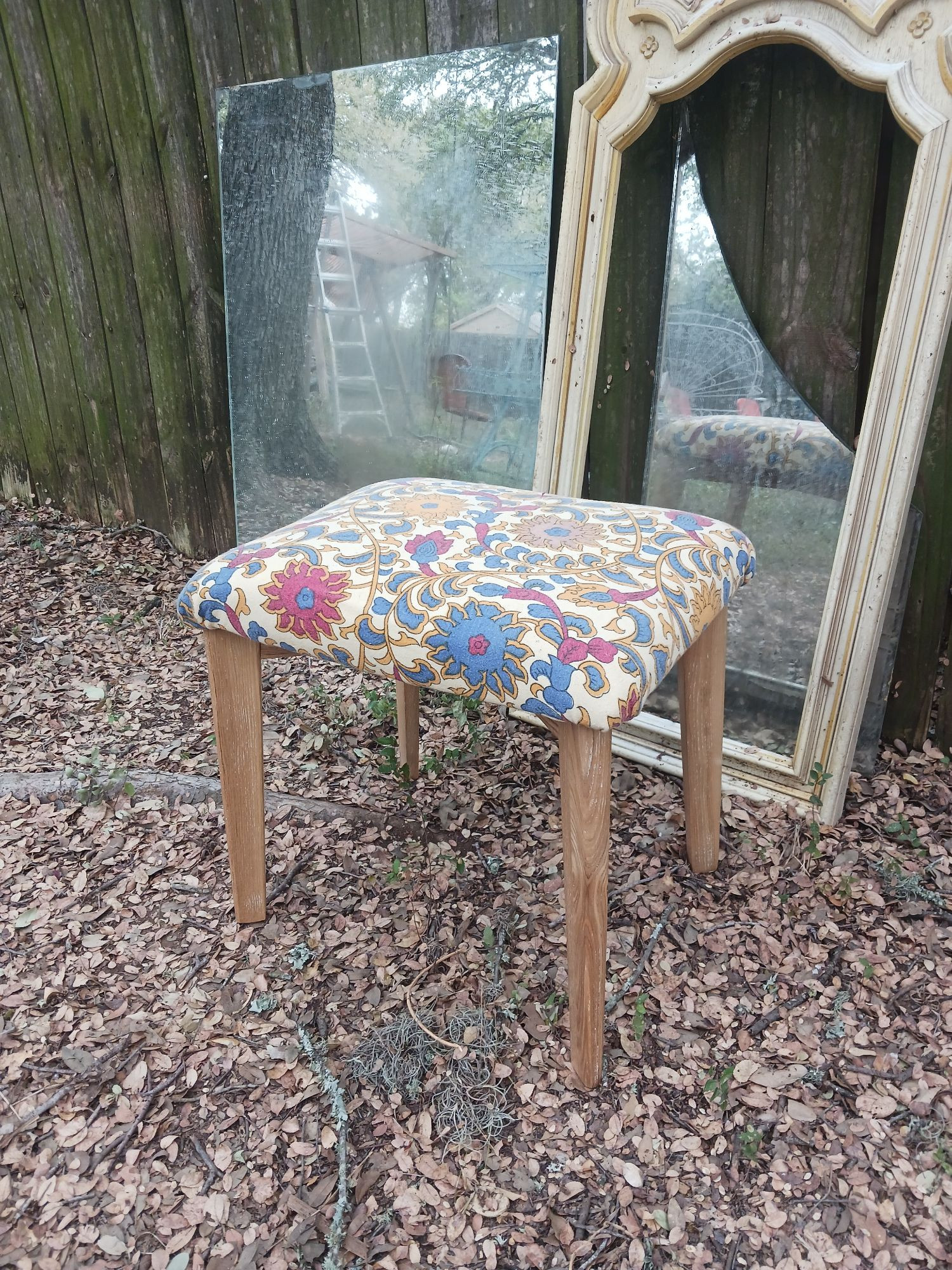 A midcentury modern blonde wood dressing stool with a colorful Indian cotton upholstery in front of a fence covered in mirrors