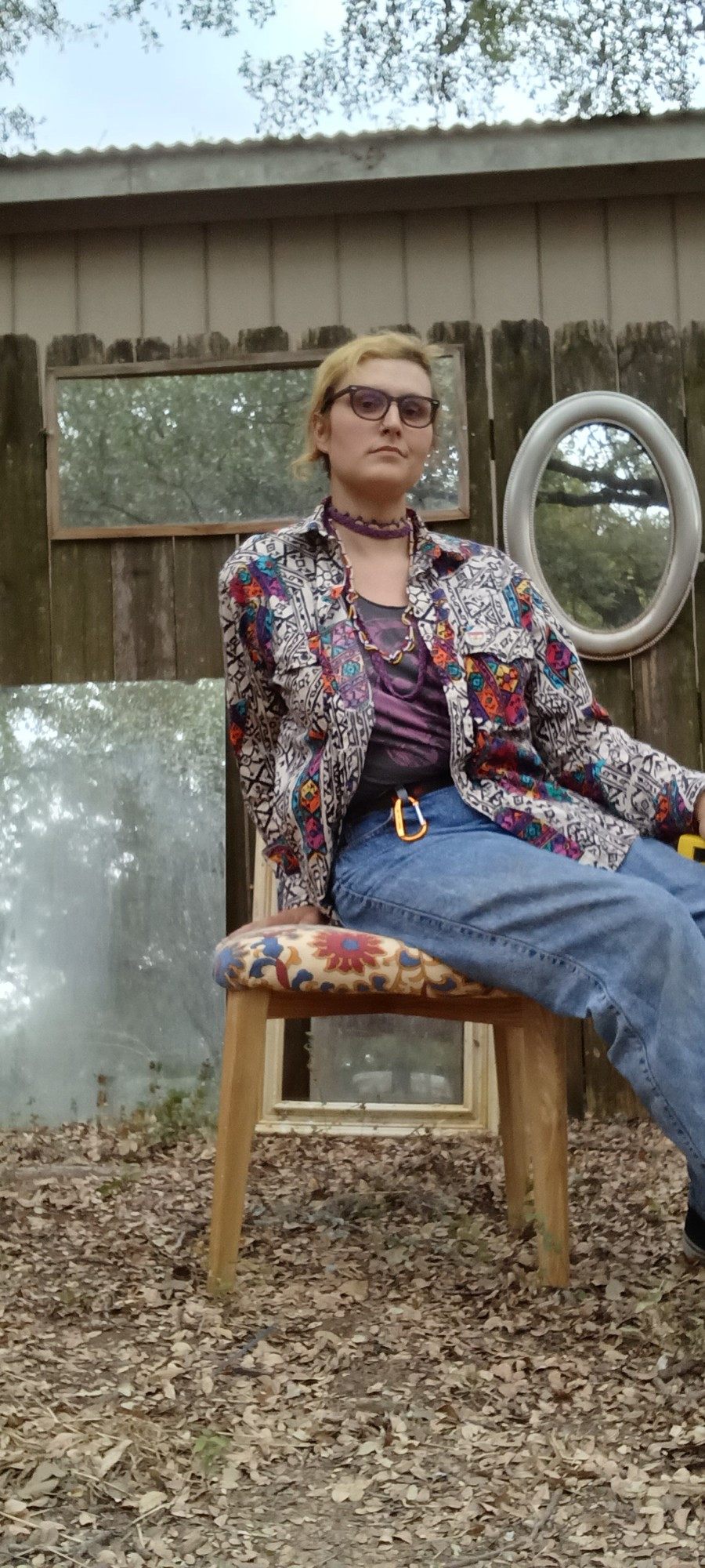 Aaron white queer sits side saddle on a midcentury modern dressing stool with yellow wood legs and colorful Indian cotton upholstery. Shey is wearing high waisted acid wash 80s mom jeans, a grey and pink tshirt, and a loudly patterned unbuttoned 80s long sleeve shirt, with multiple beaded necklaces on top. Shey is not good at smiling while modeling. A carnation colored carabiner is very obvious on sheer belt loop 