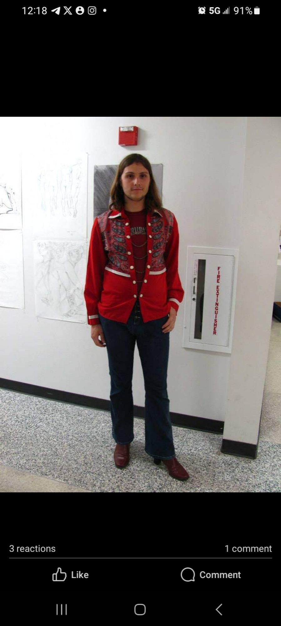 A pretransition photo of a white queer with long hair standing in an art building hallway wearing women's bell bottoms, boots, and a vintage  tshirt underneath a red abd white patterned blouse with a paliacate like pattern. The blouse doesn't close, and instead hangs open with gold chains on white buttons.