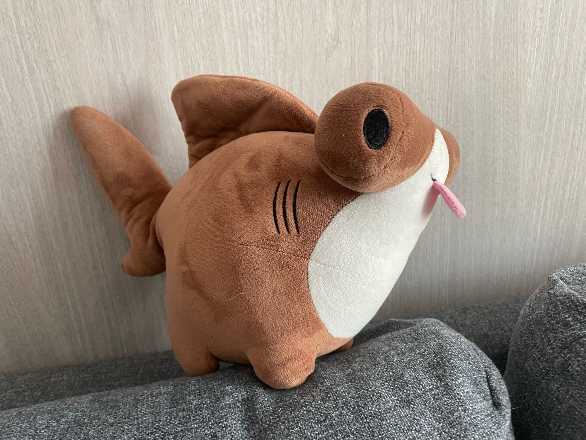 A round plush of a cartoonishly cute hammerhead shark with stubby little legs and a tongue sticking out in a blep