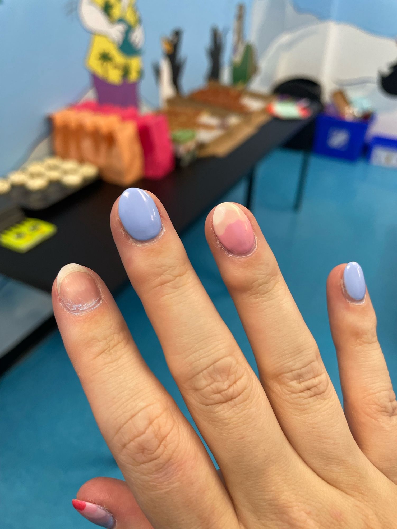 My hand, with 4 painted nails and one mostly-bare one where my acrylic dip overlay popped off. In the background is a blurry kid’s party table.