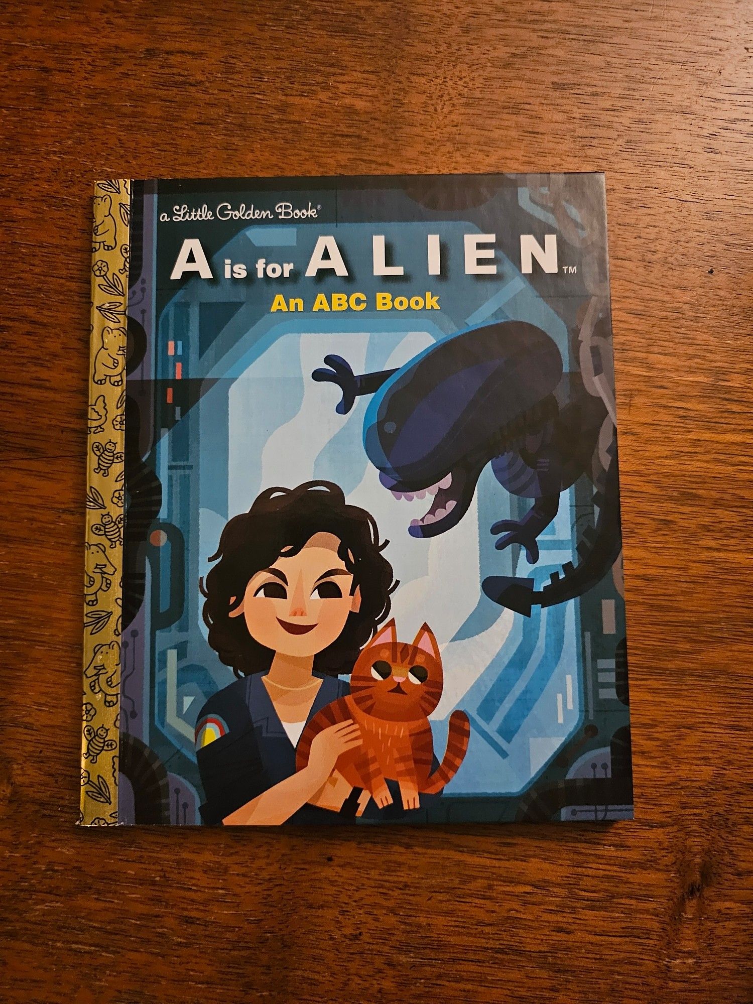 Small children's book with golden spine called "A is for Alien: An ABC Book"

Cover has Ripley holding her cat Jones. Background is a generic sci-fi space ship door. On the right a little more than half way up is a xenomorph trying to be scary