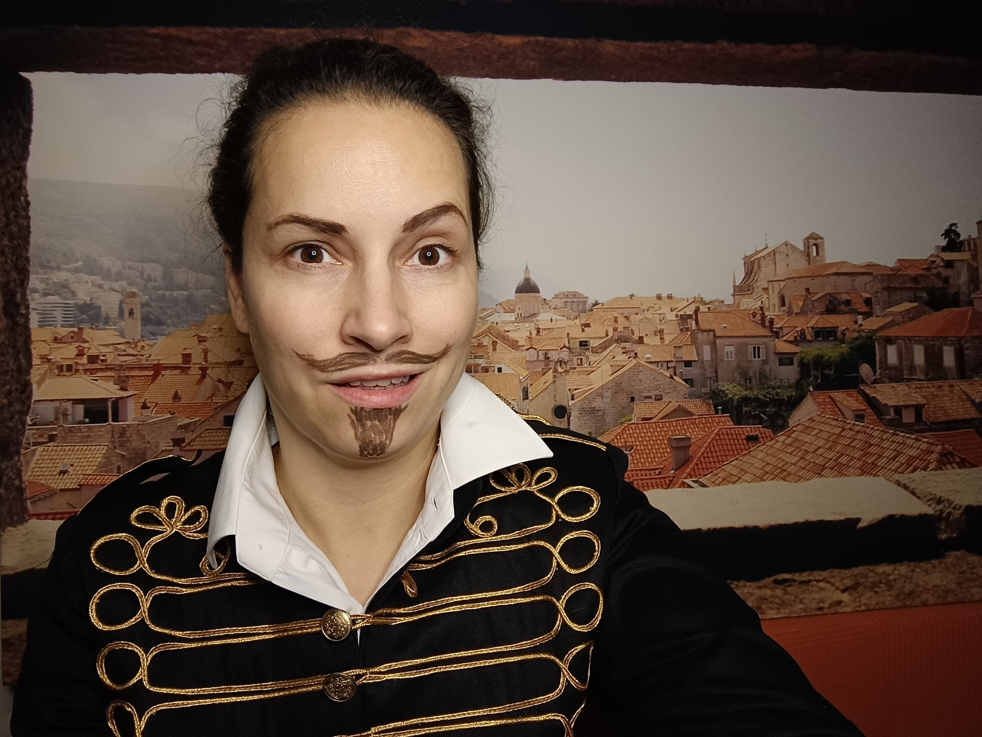 Picture of me - a white woman with dark eyes and hair pulled back, a mustache and a goatee drawn with a brown pencil, one thick eyebrow arched. I'm wearing an ornate black and gold jacket with a white shirt underneath. Background shows the roofs of Dubrovnik 