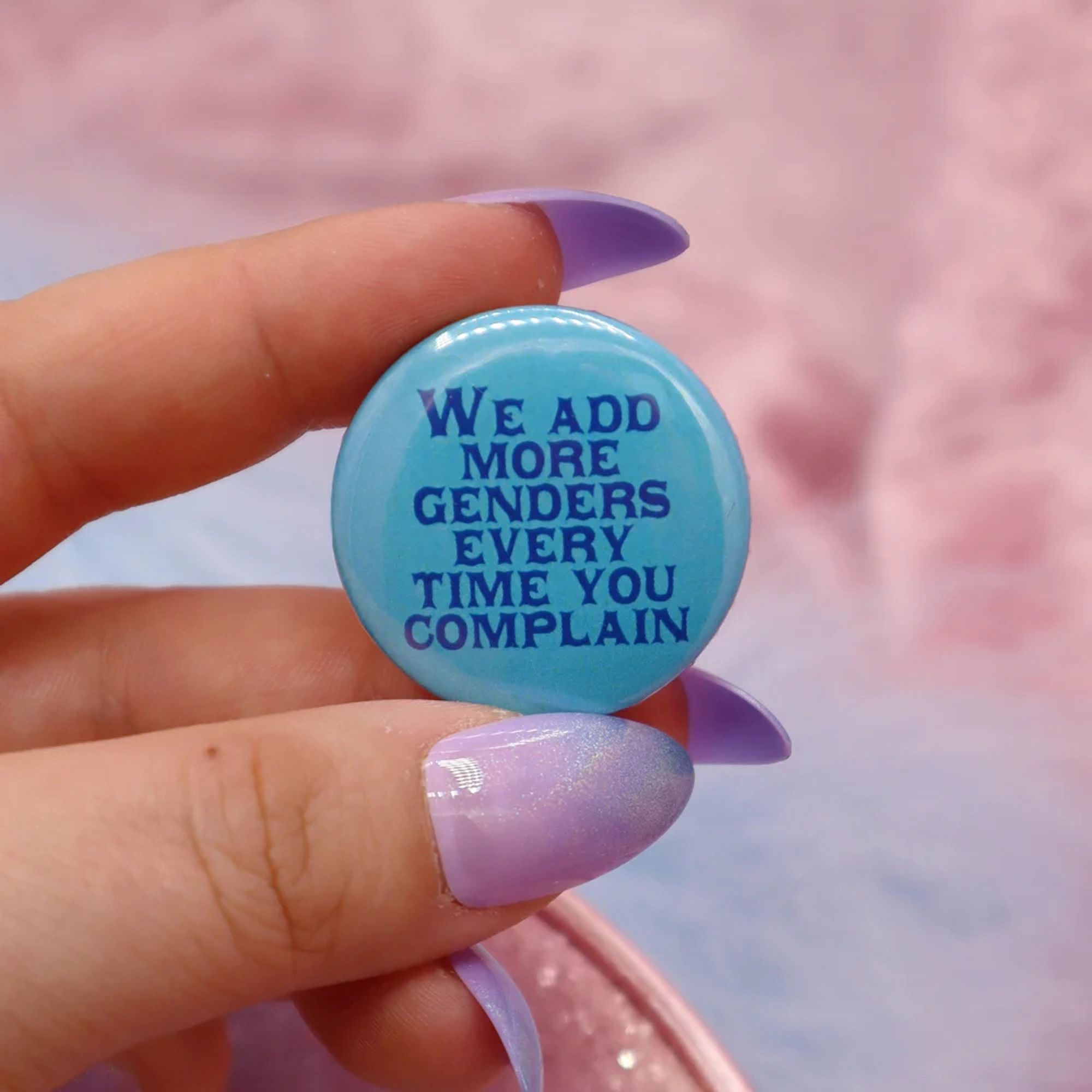 A manicured hand holding a round blue button that says "We add more genders every time you complain"