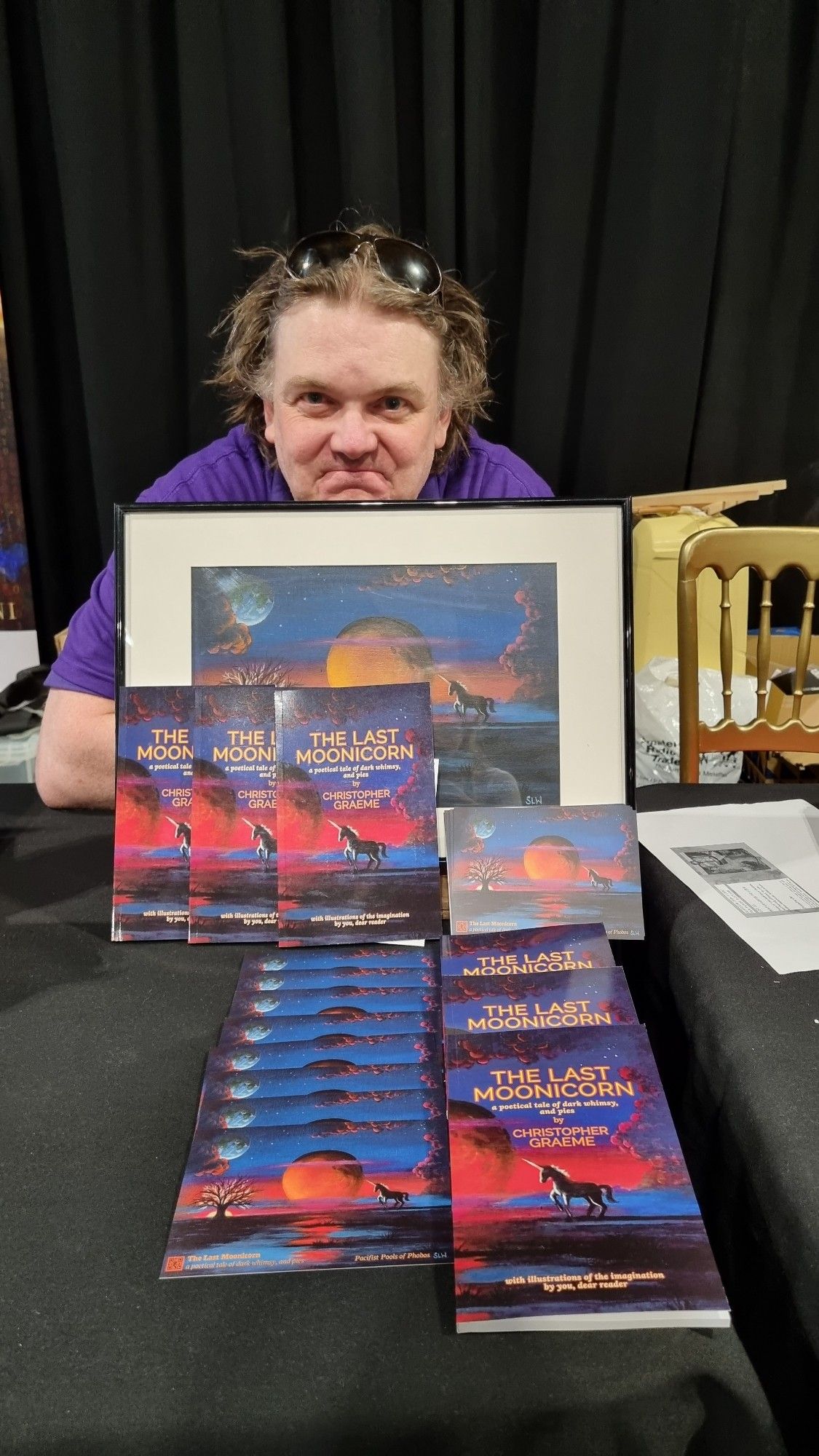 Chris behind his Worldcon book stand, with copies of The Last Moonicorn, postcards, and the cover artwork.