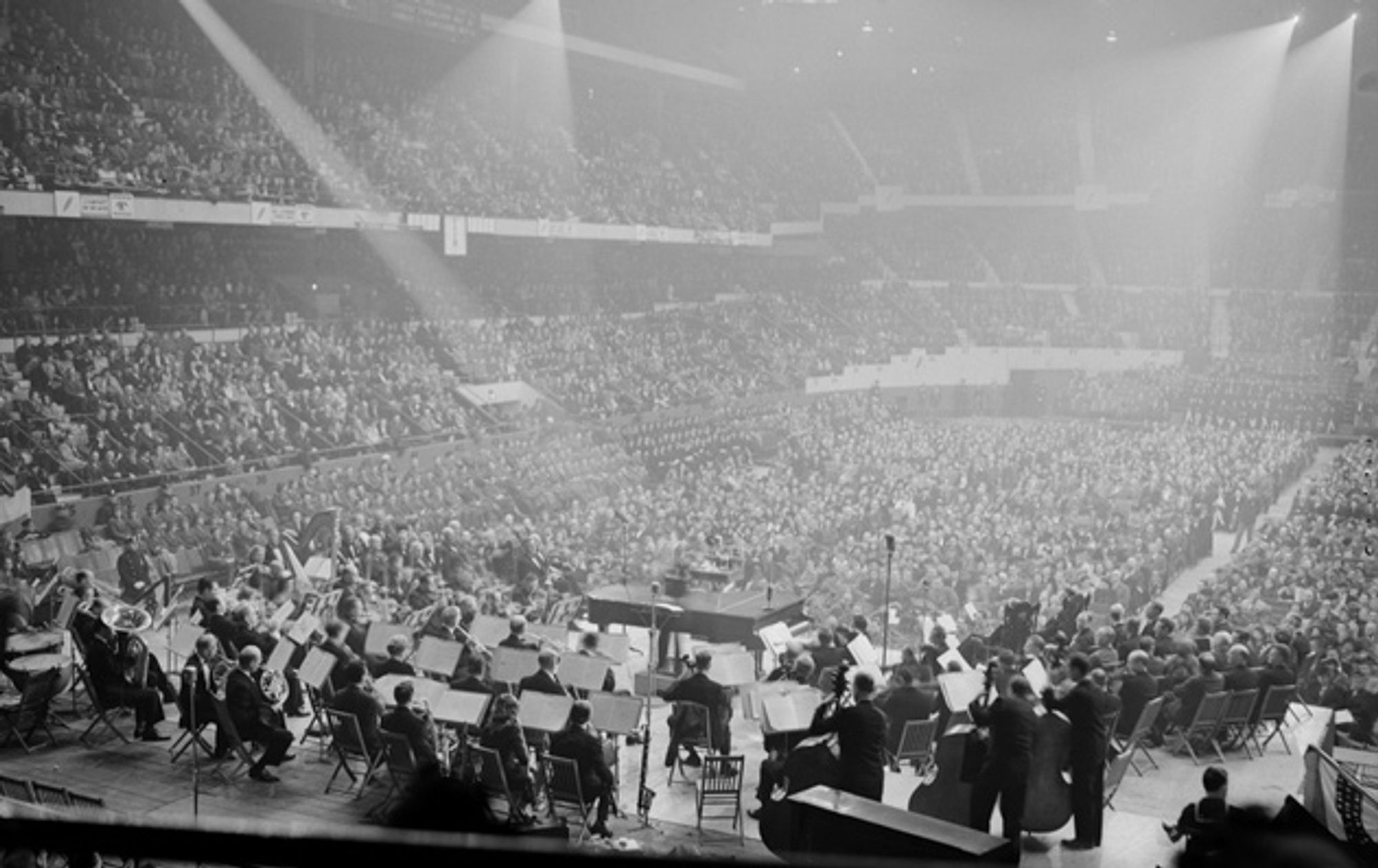 A hazy arena full of people, with an orchestra performing on a raised stage.