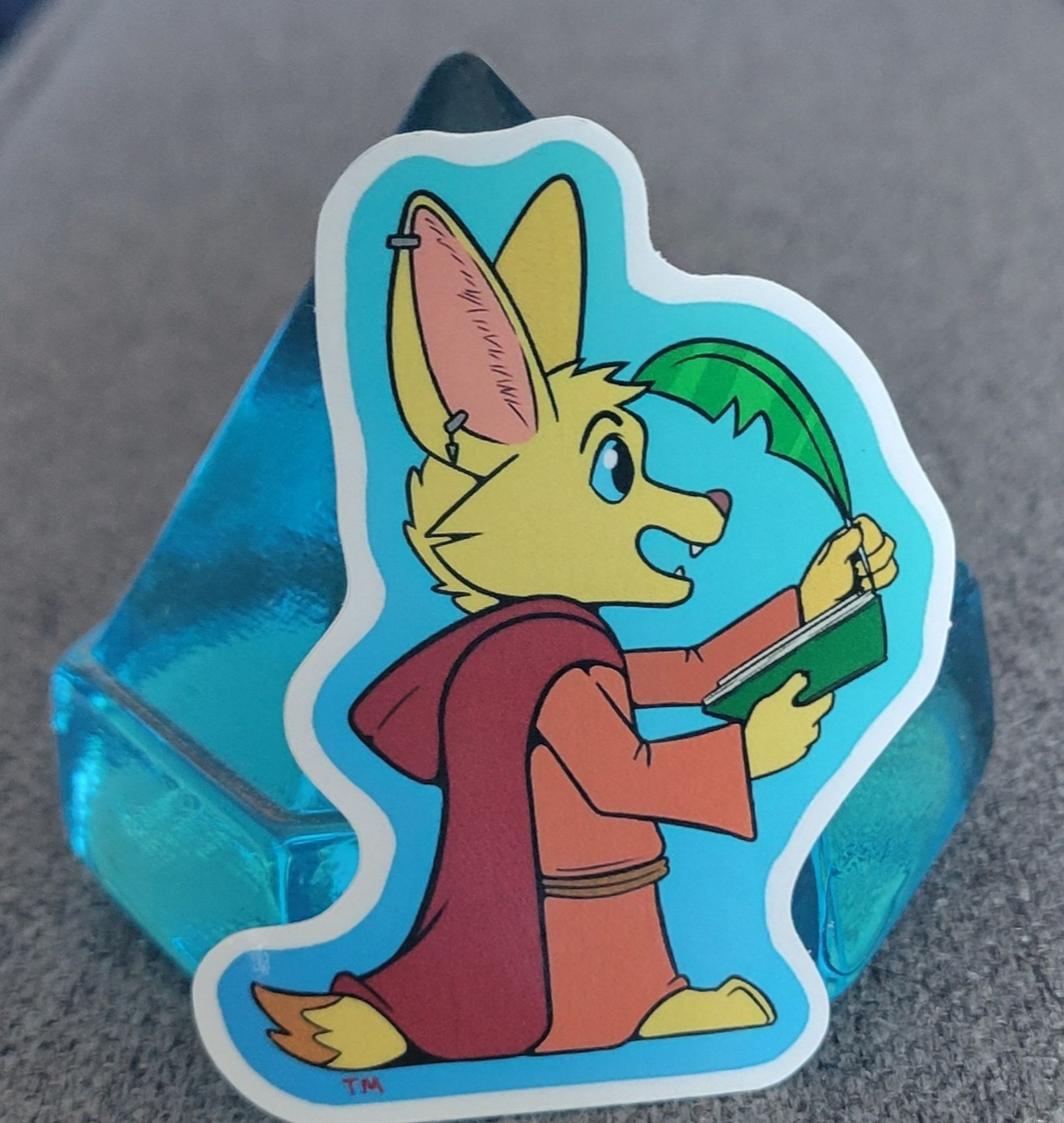 An anthropomorphic fennec in a cute chibiish style wearing a red robe with a darker read hooded cloak. They are holding a green book and quill