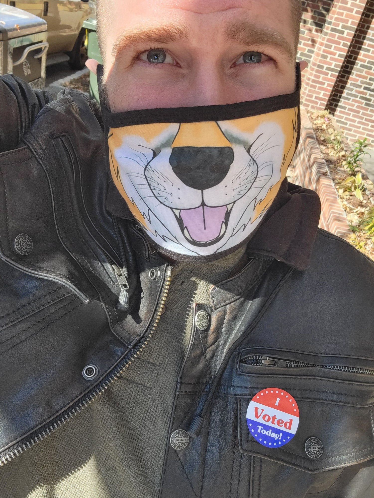 A Caucasian man with blue eyes  in a leather jacket wearing a face mask in the shape of a fox muzzle. The jacket is adorned with a sticker that says "I voted" 