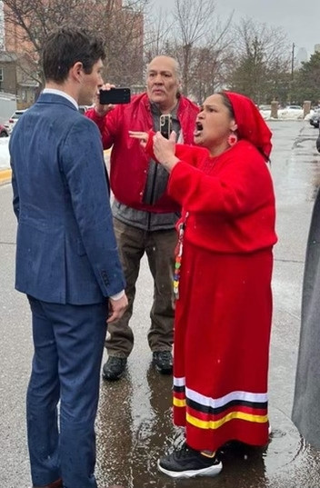 Nicole Mason, wearing all red and a ribbon skirt with the 4 sacred colors, shouting angrily at Mayor Frey while another community member films on his phone. Mayor Frey, as always, looks vapid and uncaring.