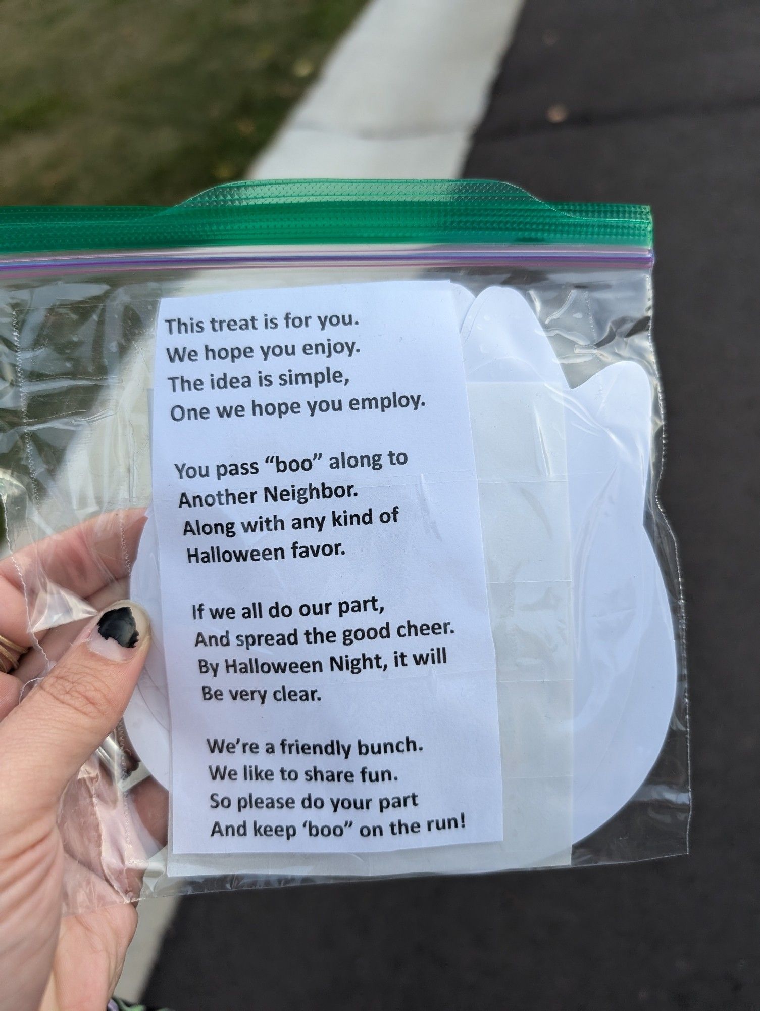 White person's hand with black shitty nail polish holding a ziplock full of ghost shaped stickers that has a poem in it: "This treat is for you, we hope you enjoy, the idea is simple, one we hope you enjoy
 You pass "boo" along to another neighbor, along with any kind of Halloween favor. If we all do our part and spread the good cheer, by Halloween night it will be very clear. We're a friendly bunch, we like to share fun, so please do your part and keep "boo" on the run!"