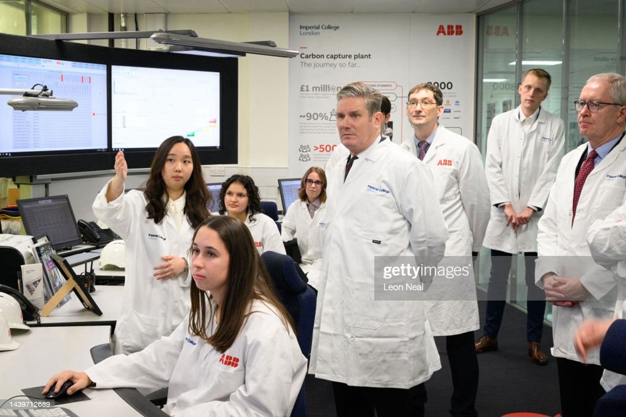 Keir Starmer in a scientist coat stood near a bunch of women sat down with men looking in the background